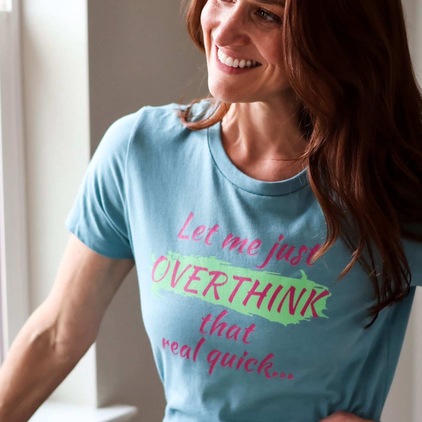 Woman smiles while wearing a blue top that reads Let me just VERTHINK that real quick in bright green and pink.