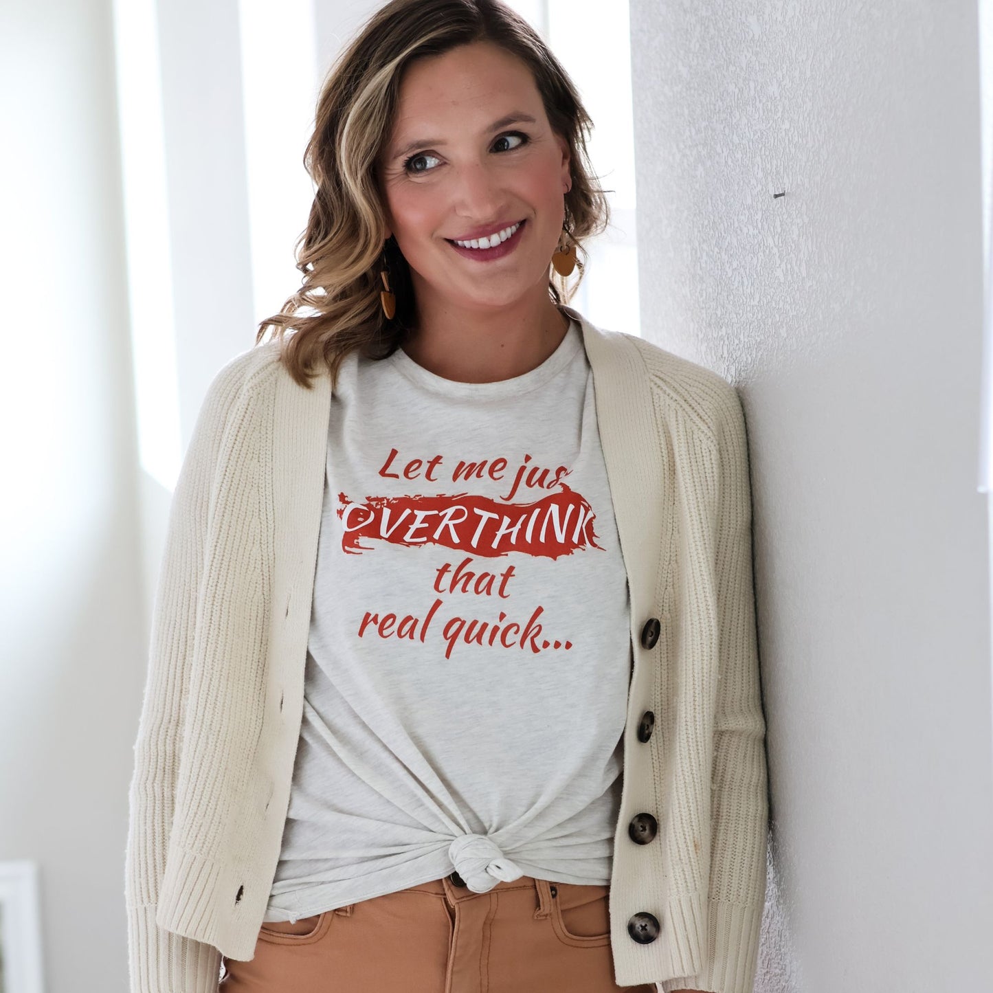 Woman wearing cream Overthinking tshirt with cardigan an dtan pants stands against a white wall.