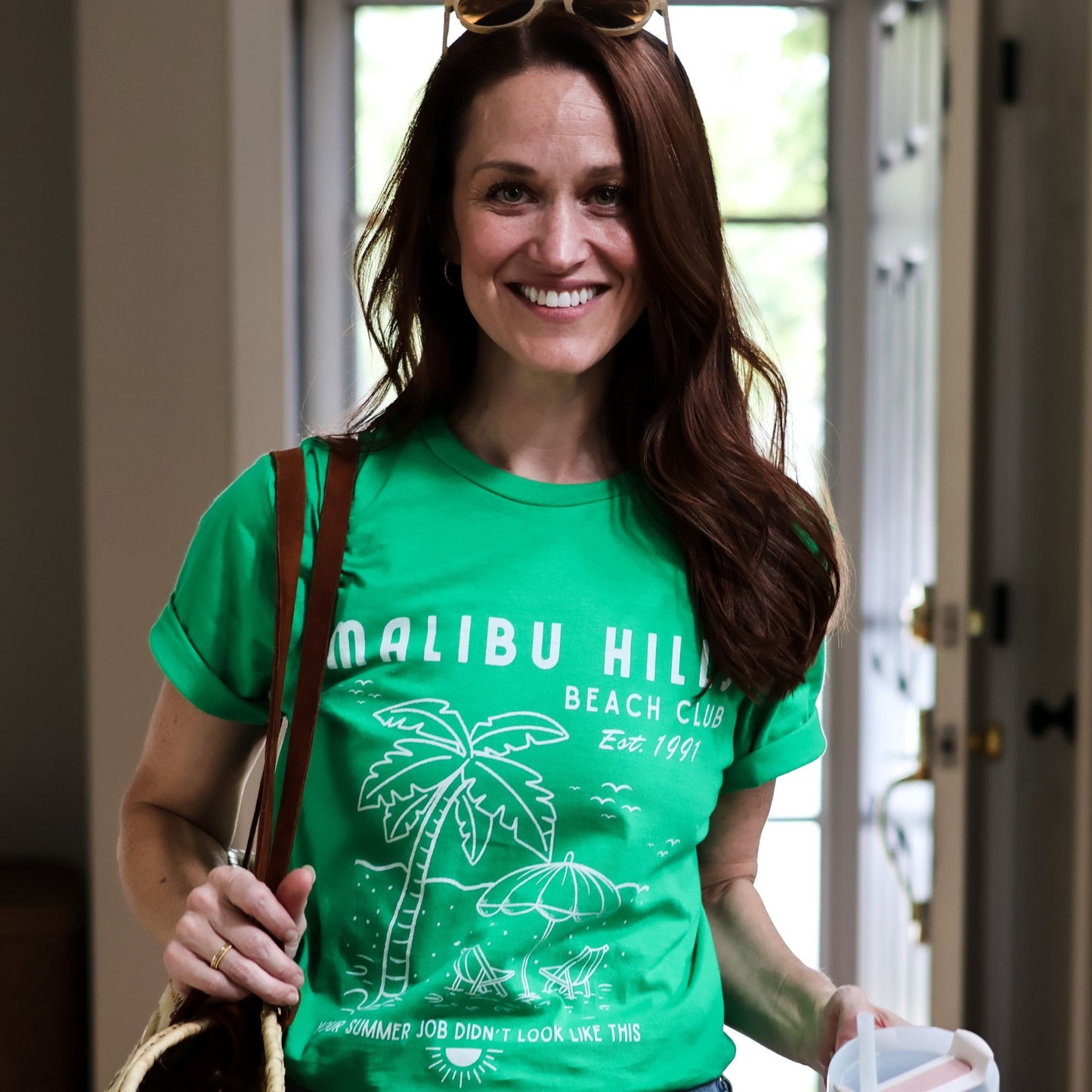 Woman wearing a green 'Malibu Hills Beach Club' t-shirt with palm tree and umbrella design.