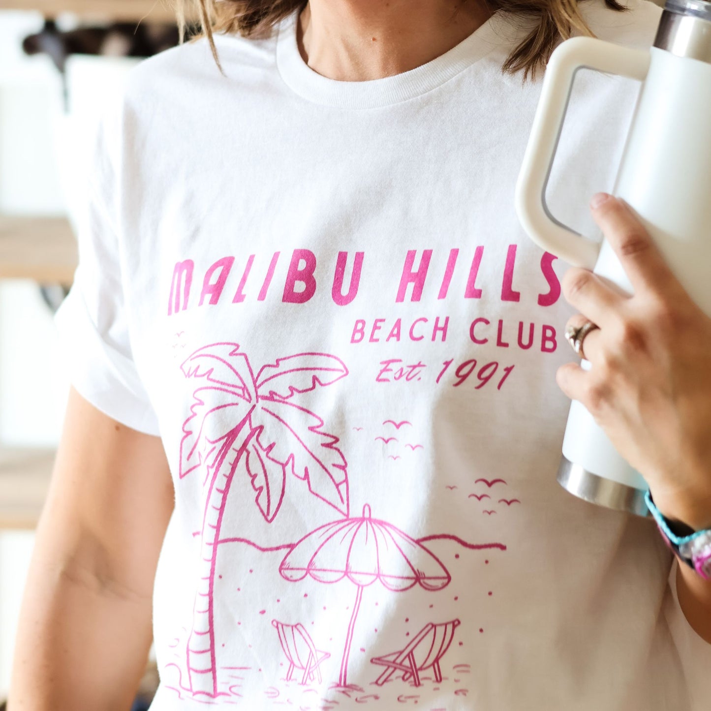 Woman wearing a white t-shirt with pink 'Malibu Hills Beach Club' design, holding a white mug.