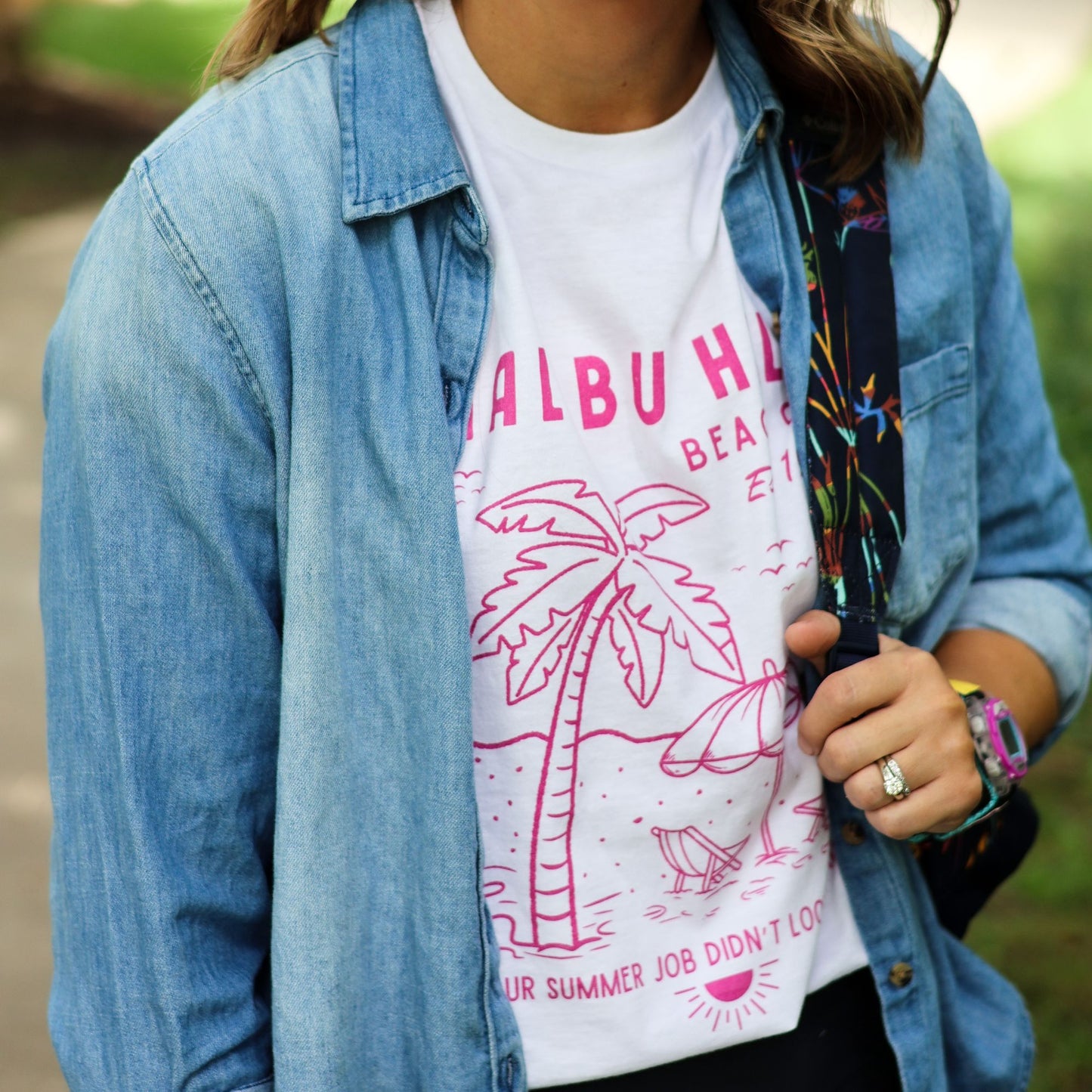 Woman outdoors wearing a white t-shirt with pink graphics and text, paired with a denim shirt and shoulder bag.