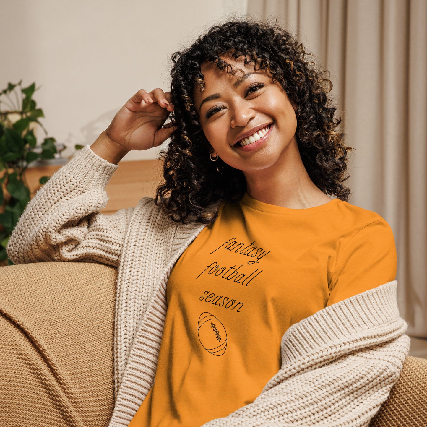 Smiling woman with curly hair sits on a couch wearing a tee shirt that reads "FANTASY FOOTBALL SEASON" along with an image of a football. The font is a thin and flowy cursive that is pretty and feminine.