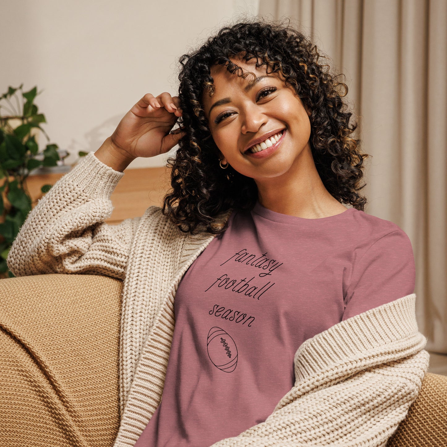 Smiling woman with curly hair sits on a couch wearing a tee shirt that reads "FANTASY FOOTBALL SEASON" along with an image of a football. The font is a thin and flowy cursive that is pretty and feminine.
