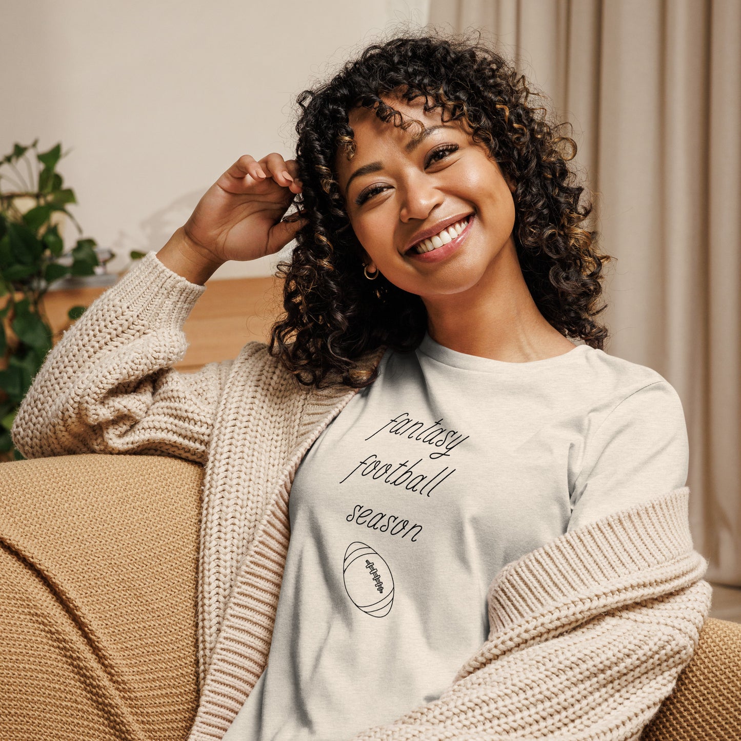 Smiling woman with curly hair sits on a couch wearing a tee shirt that reads "FANTASY FOOTBALL SEASON" along with an image of a football. The font is a thin and flowy cursive that is pretty and feminine.