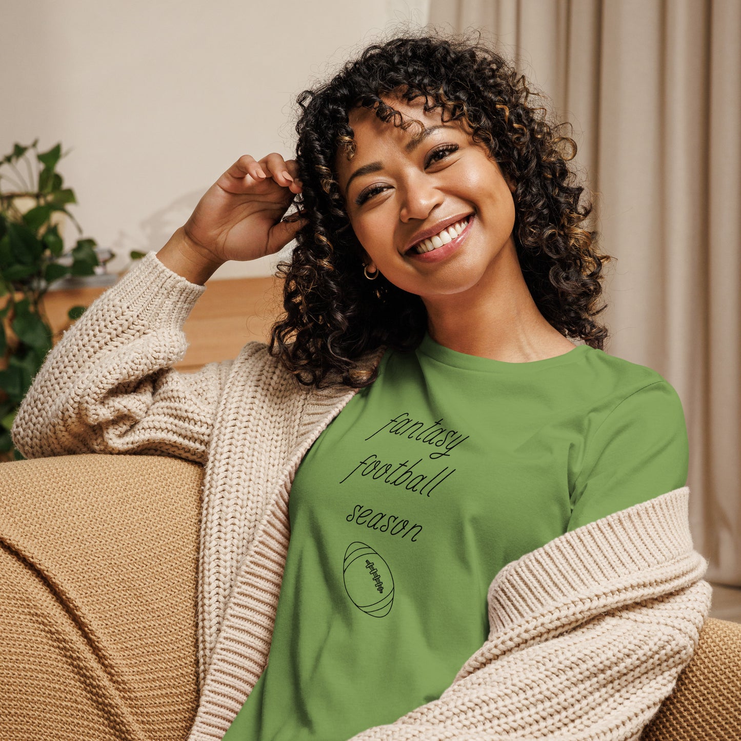 Smiling woman with curly hair sits on a couch wearing a tee shirt that reads "FANTASY FOOTBALL SEASON" along with an image of a football. The font is a thin and flowy cursive that is pretty and feminine.