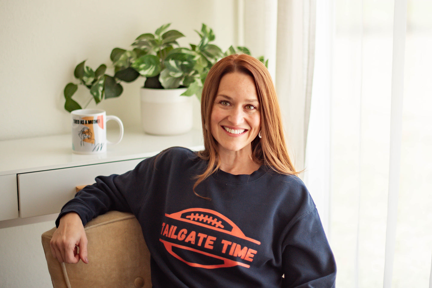 Woman looks cozy in a navy blue sweatshirt with an large football graphic and the words "Tailgate time" in distressed letting.  The blue sweatshirt and bright orange font in perfect for chicago bears fans, denver broncos fans, or university of illinois fans.