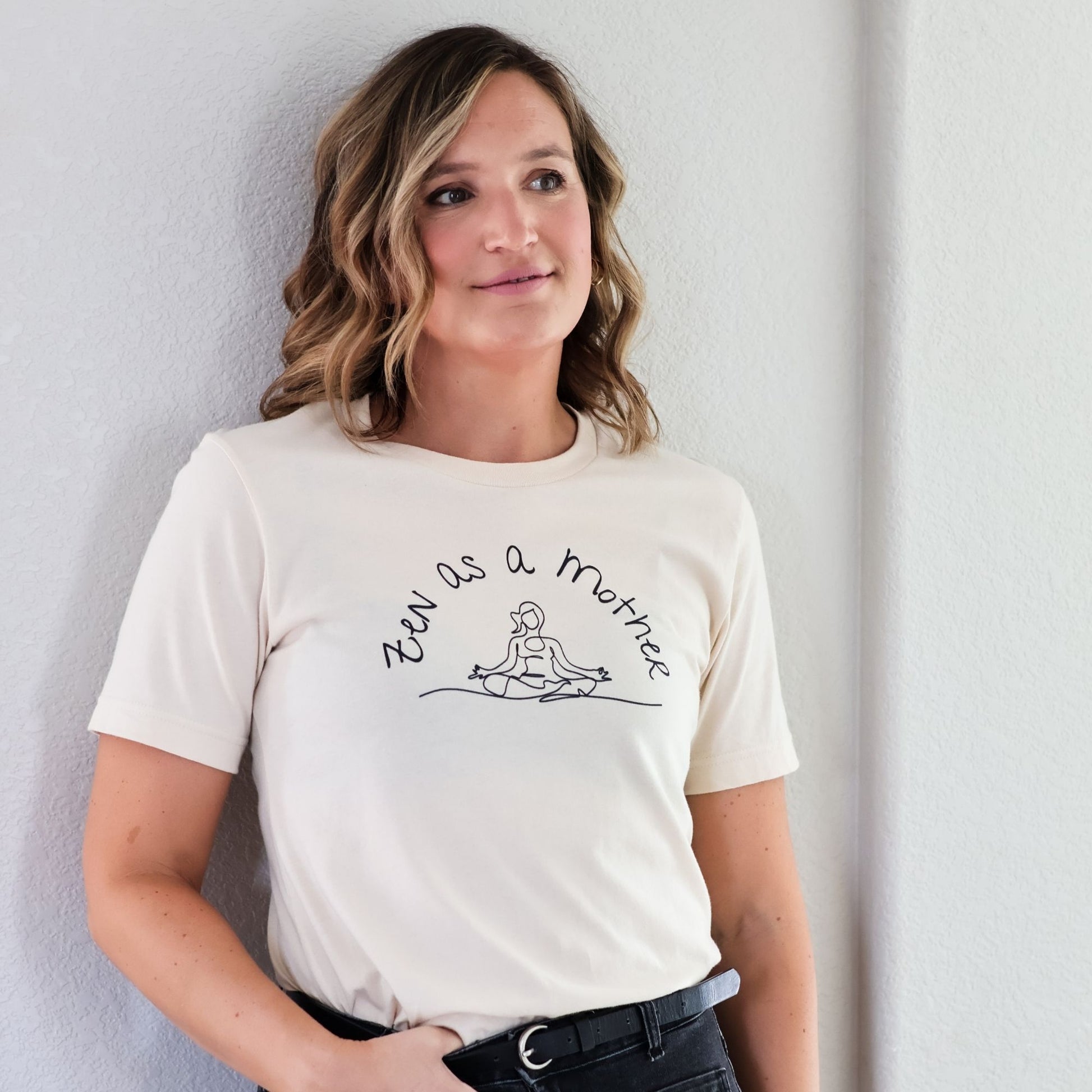 Woman leans against wall wearing Zen As A Mother t-shirt in cream with black jeans