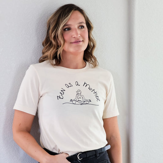 Woman leans against wall wearing Zen As A Mother t-shirt in cream with black jeans