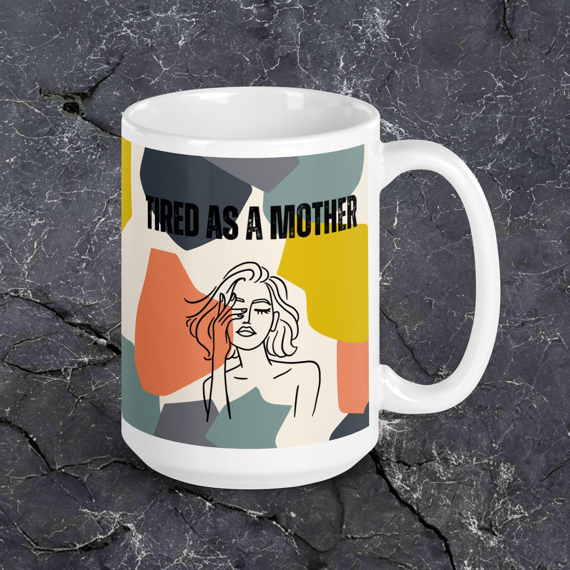 A white ceramic mug featuring the text "TIRED AS A MOTHER" and an abstract illustration of a weary woman with tousled hair sits on a black granite background. The design incorporates a modern, colorful geometric background with shapes in yellow, orange, gray, and teal.
