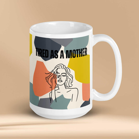 A white ceramic mug featuring the text "TIRED AS A MOTHER" and an abstract illustration of a weary woman with tousled hair. The design incorporates a modern, colorful geometric background with shapes in yellow, orange, gray, and teal.