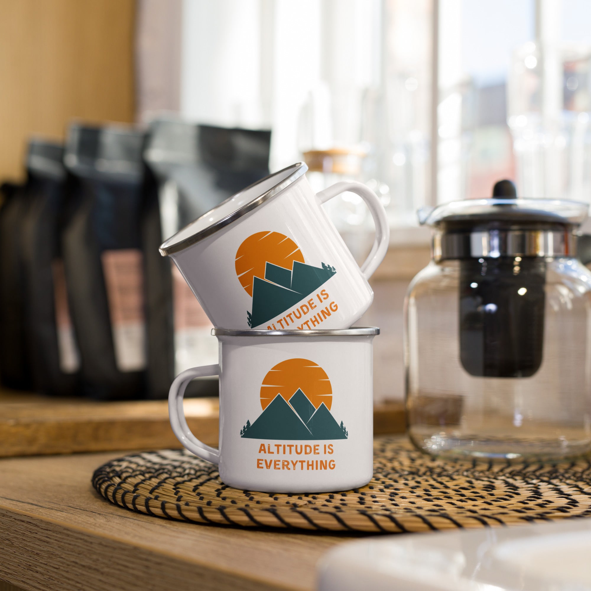 Two white enamel mugs stacked on a woven placemat, displaying "ALTITUDE IS EVERYTHING" with a mountain and sun graphic. The mugs are shown in a kitchen setting with a French press nearby, emphasizing their use for coffee or tea.