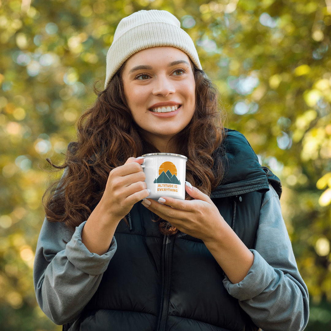 Woman with puffy vest and beanie hat enjoys warm beverage in a white enamel mug. The camping mug reads "altitude is everything" alongside a graphic of mountains.