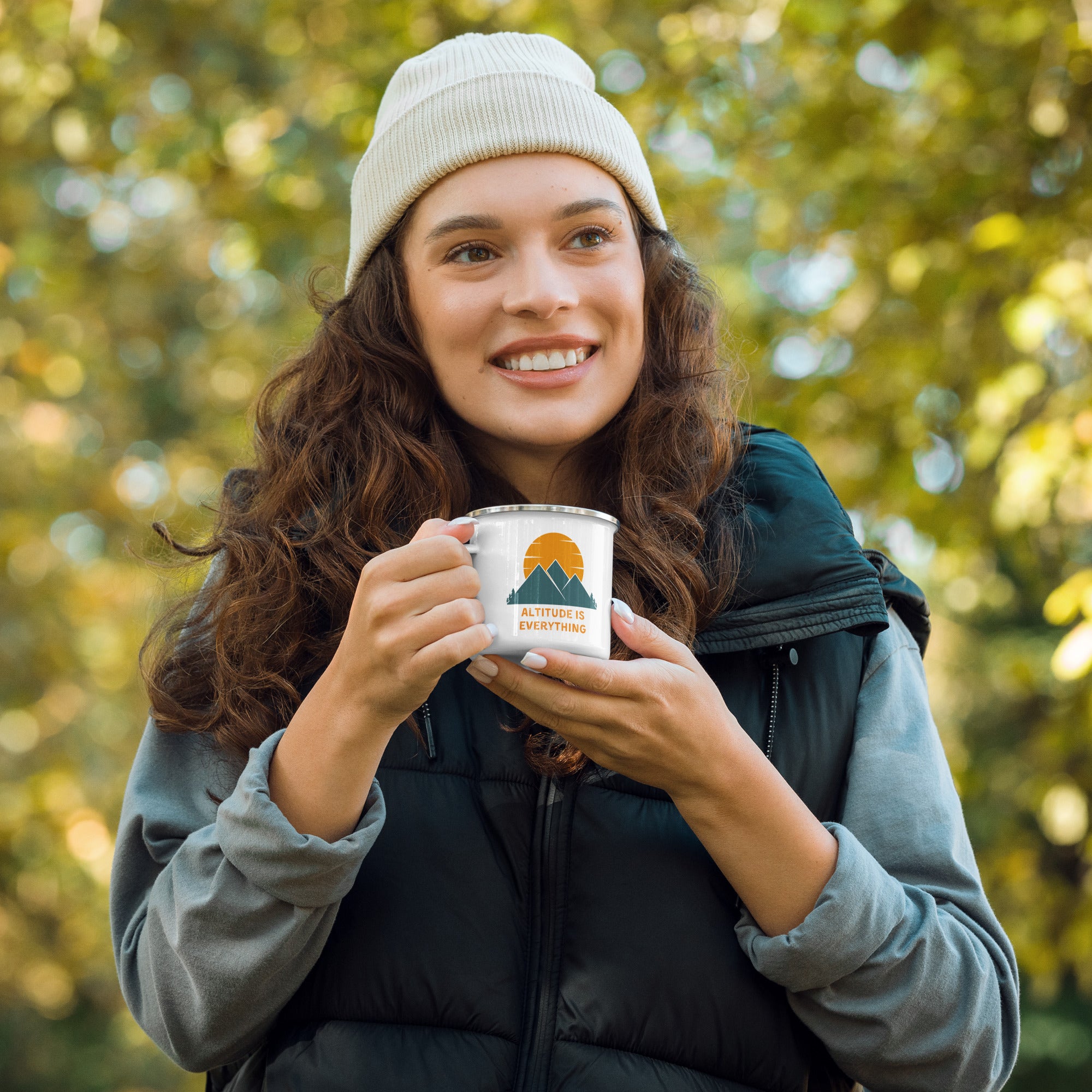 Woman with puffy vest and beanie hat enjoys warm beverage in a white enamel mug. The camping mug reads "altitude is everything" alongside a graphic of mountains.