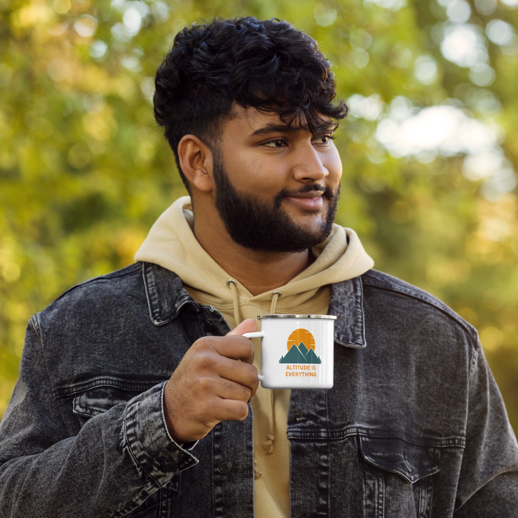 Bearded man dressed in hooded sweatshirt and jacket enjoys beverage outdoors from a white camping mug. ENamel mug says "altitude is everything" in orange. It's accompanied by a graphic of mountains and a sun.