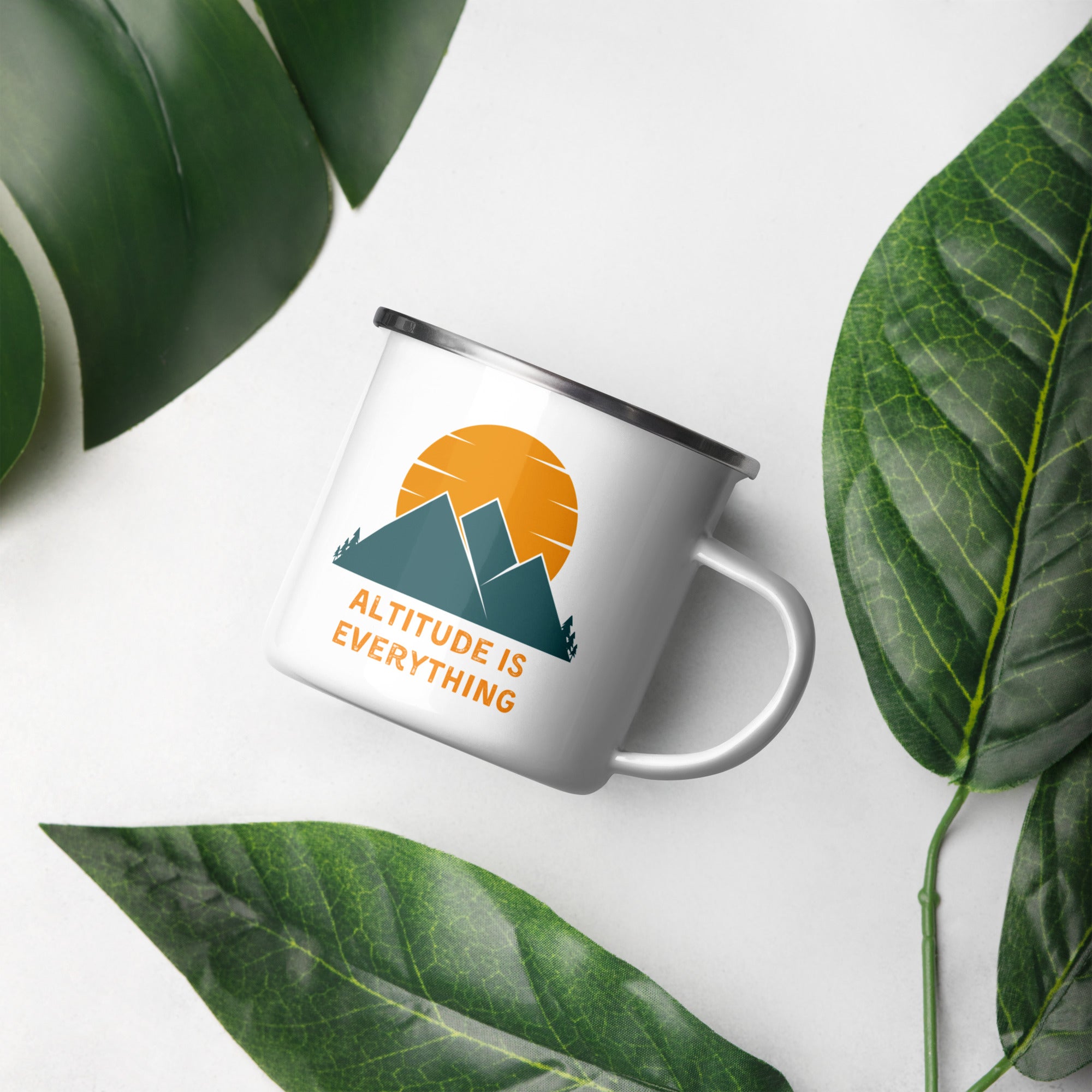 White enamel camping mug sits next to a leafy plant. The mug reads "altitude is everything" and displays a sun and mountain design.