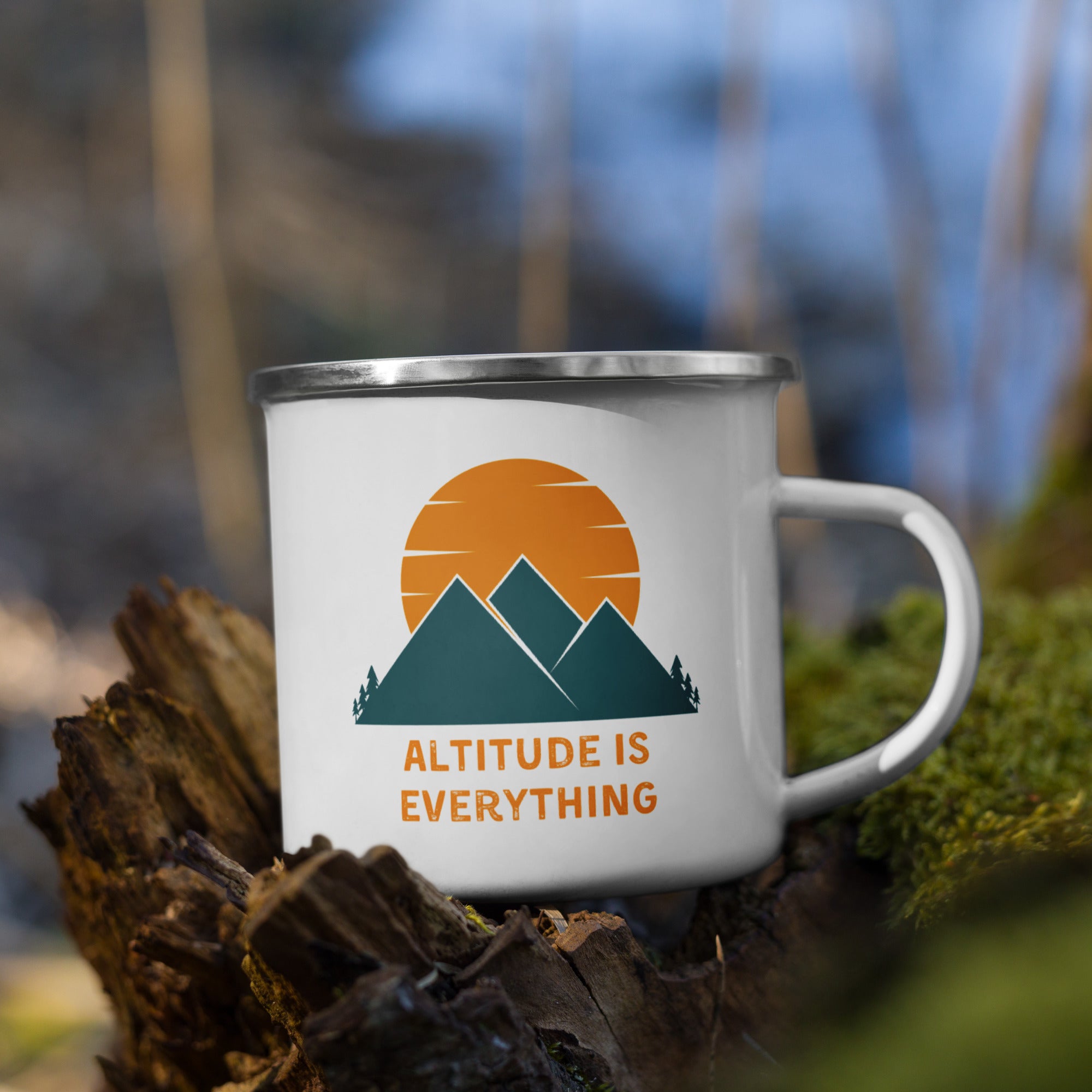 White enamel camping mug featuring a graphic of mountains and sun with the text "ALTITUDE IS EVERYTHING" in orange. The mug is photographed outdoors in nature on top of bark and moss.