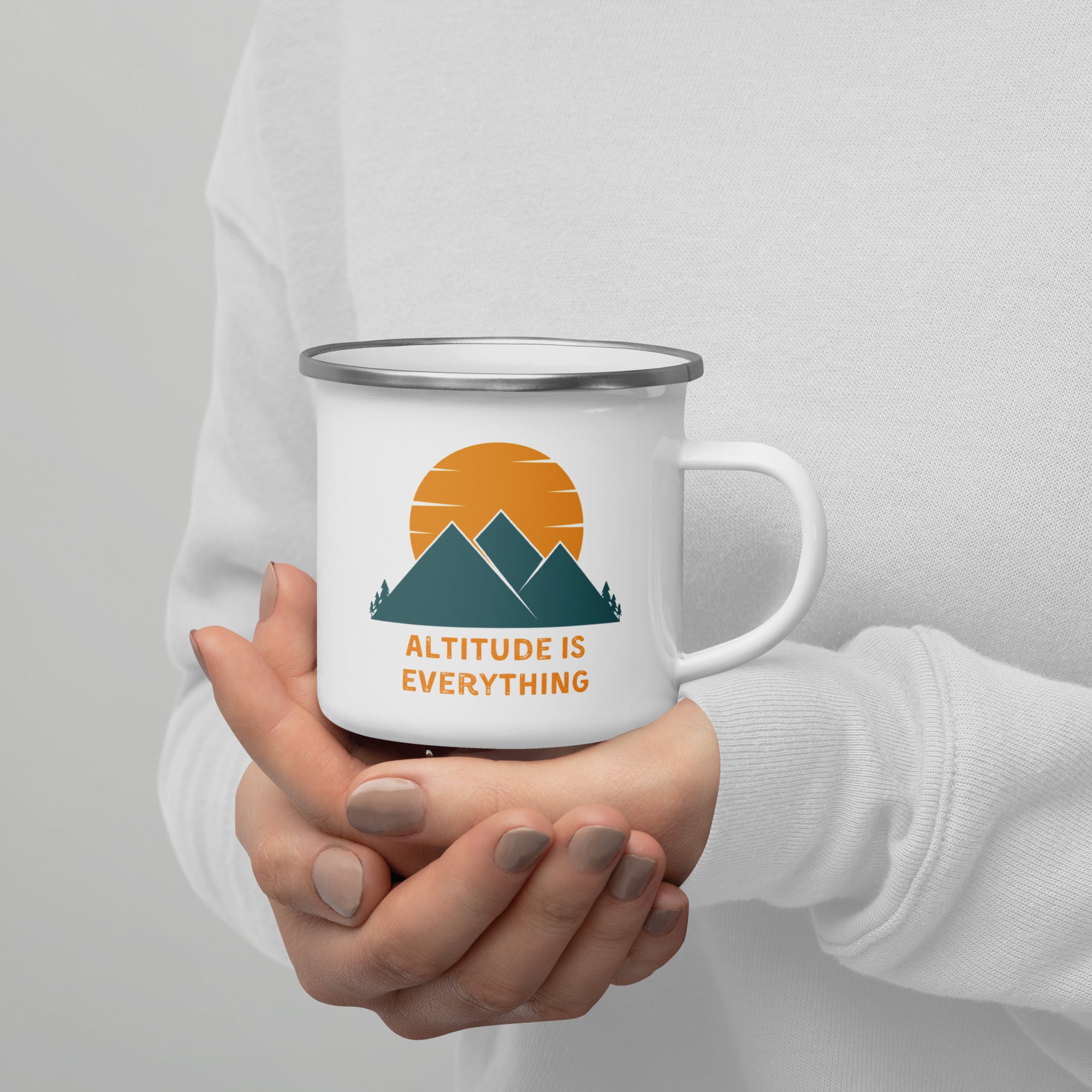 Person in a white sweatshirt cups a white enamel camping mug that says "altitude is everything" with a mountain and sun scenic design.