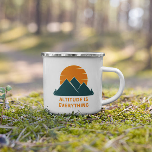 White enamel camping mug featuring a graphic of mountains and sun with the text "ALTITUDE IS EVERYTHING" in orange. The mug is photographed outdoors on mossy ground with a blurred forest background.