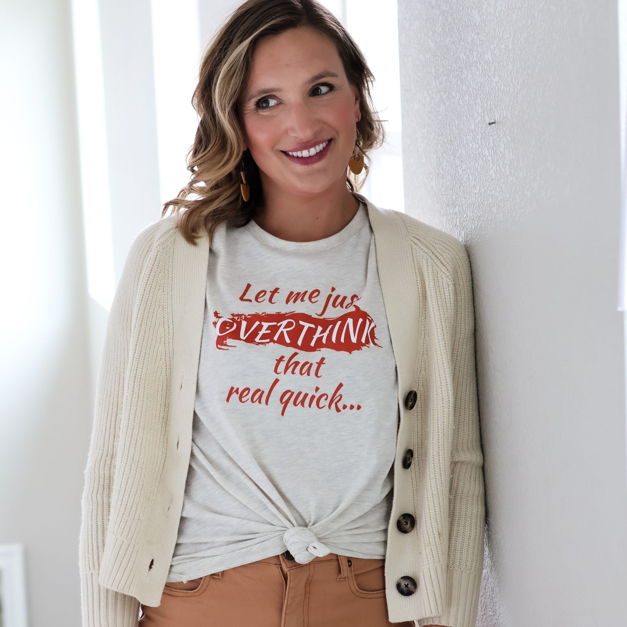 Woman wearing cream Overthinking tshirt with cardigan an dtan pants stands against a white wall.