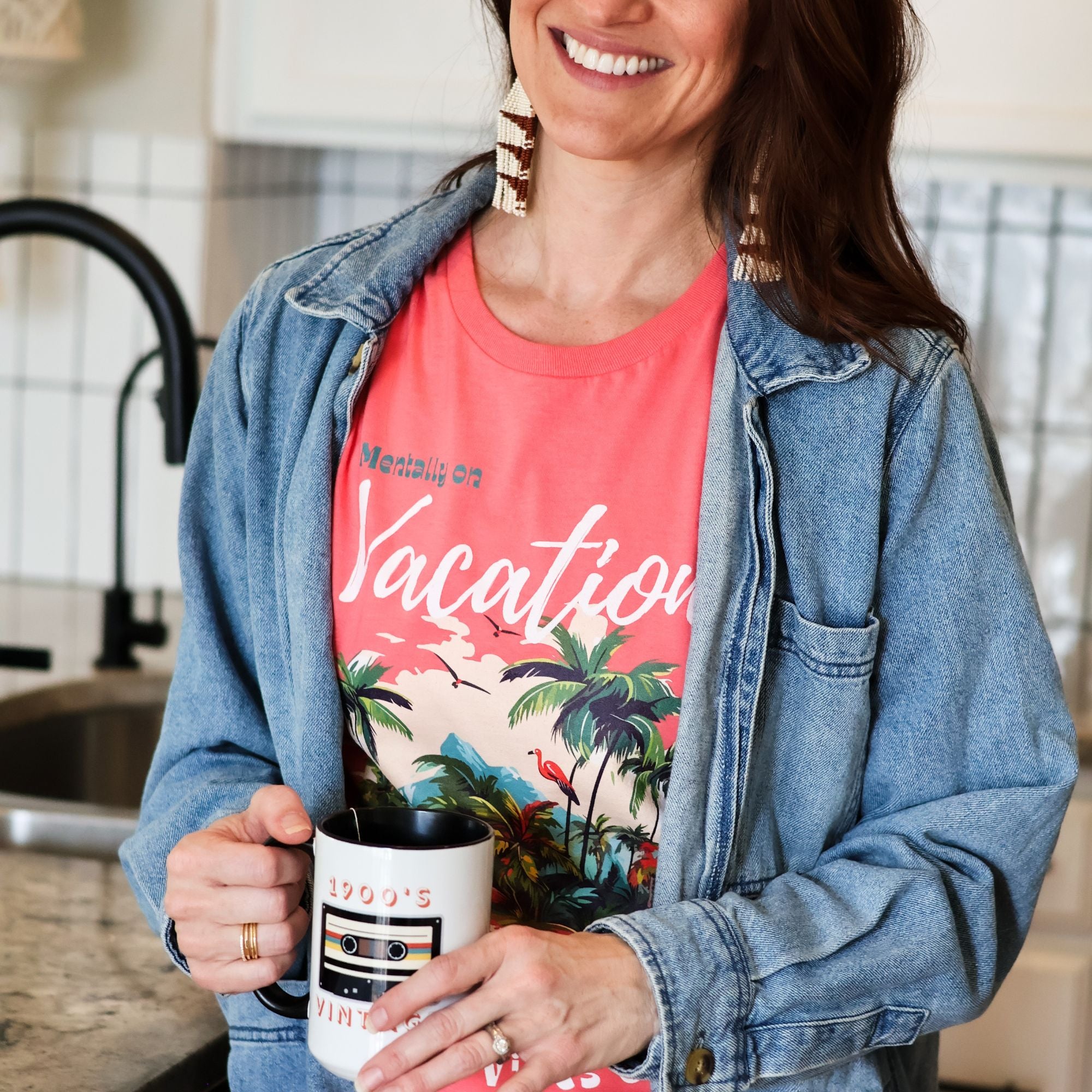 Woman holding a mug in a kitchen, wearing a tropical graphic tee that reads 'Mentally on vacation'  design with vibrant island graphic