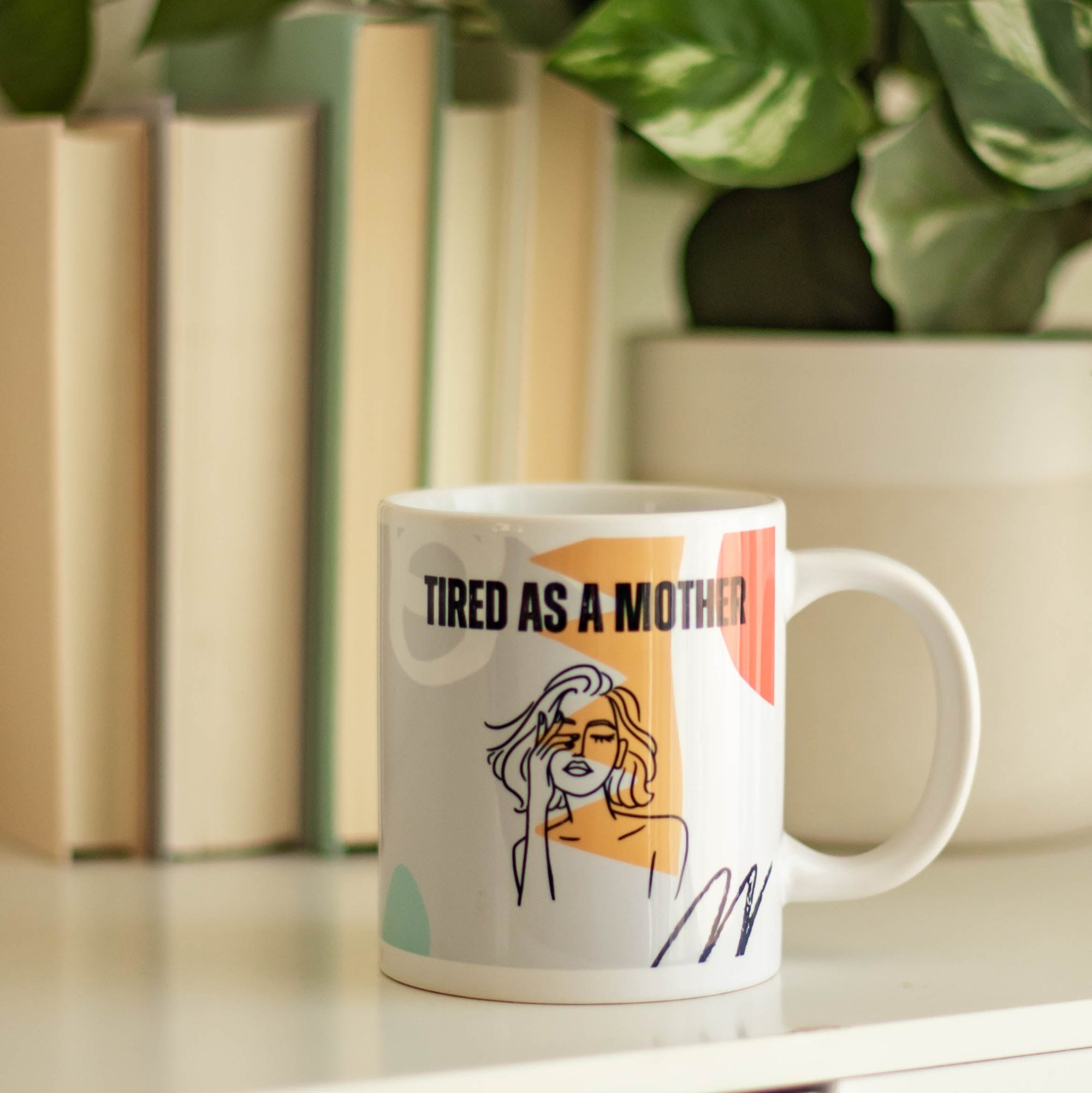 Colorful Mug with Tired as a Mother text and deisgn on a surface with books and plants in the background