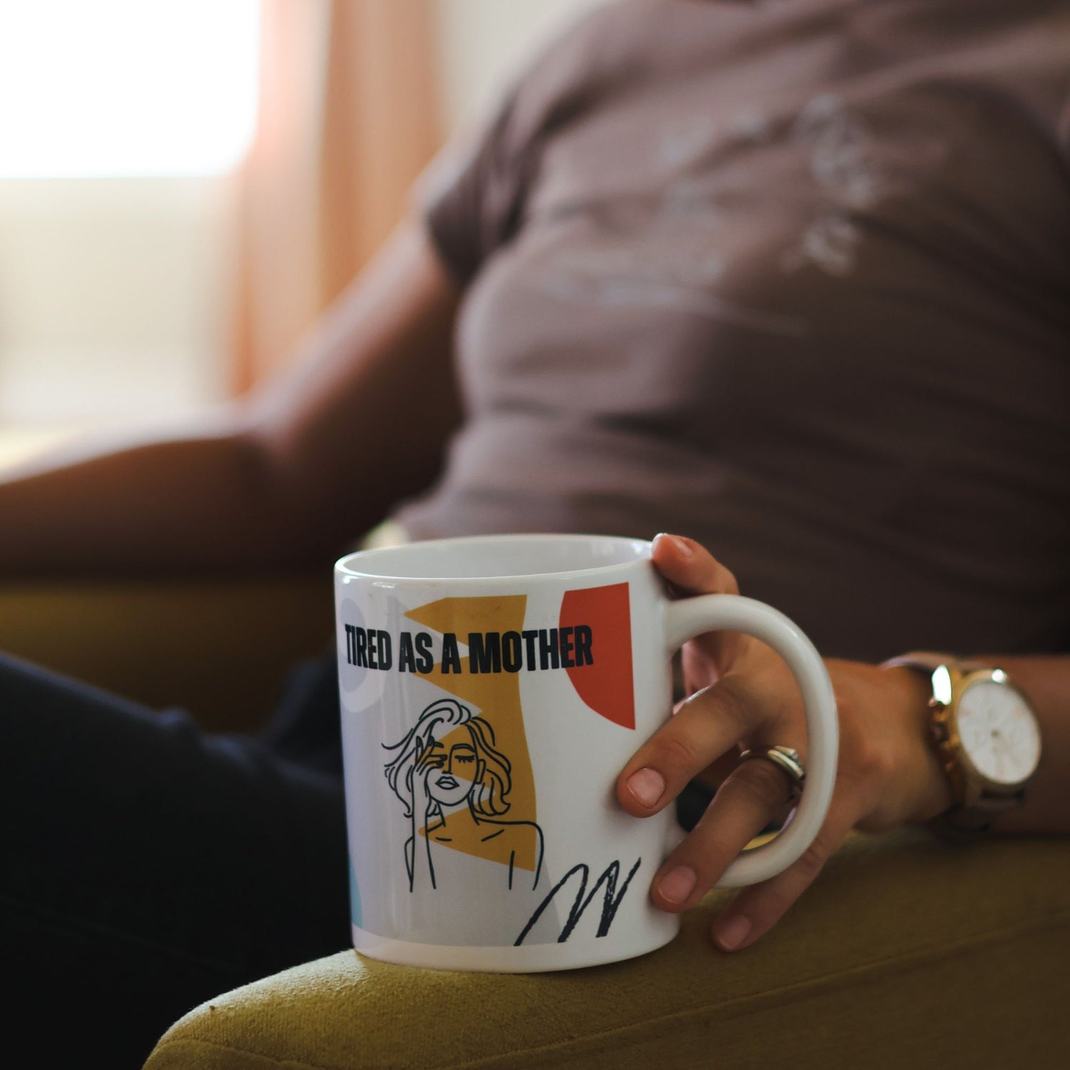 Woman holding a mug with Tired as a Mother text and female graphics, sitting on a couch.