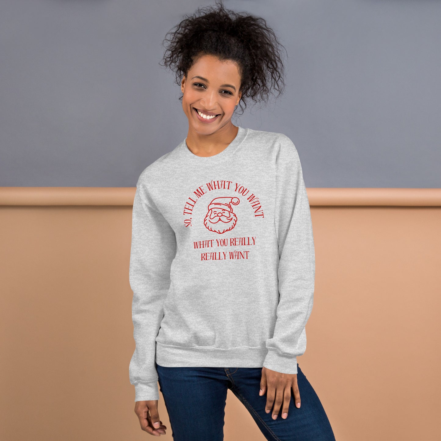 Smiling woman wears a red and gray crewneck sweatshirt featuring Santa's face and text that reads "So Tell Me What You Want What You Really Really Want," playfully referencing the Spice Girls song while incorporating holiday humor.