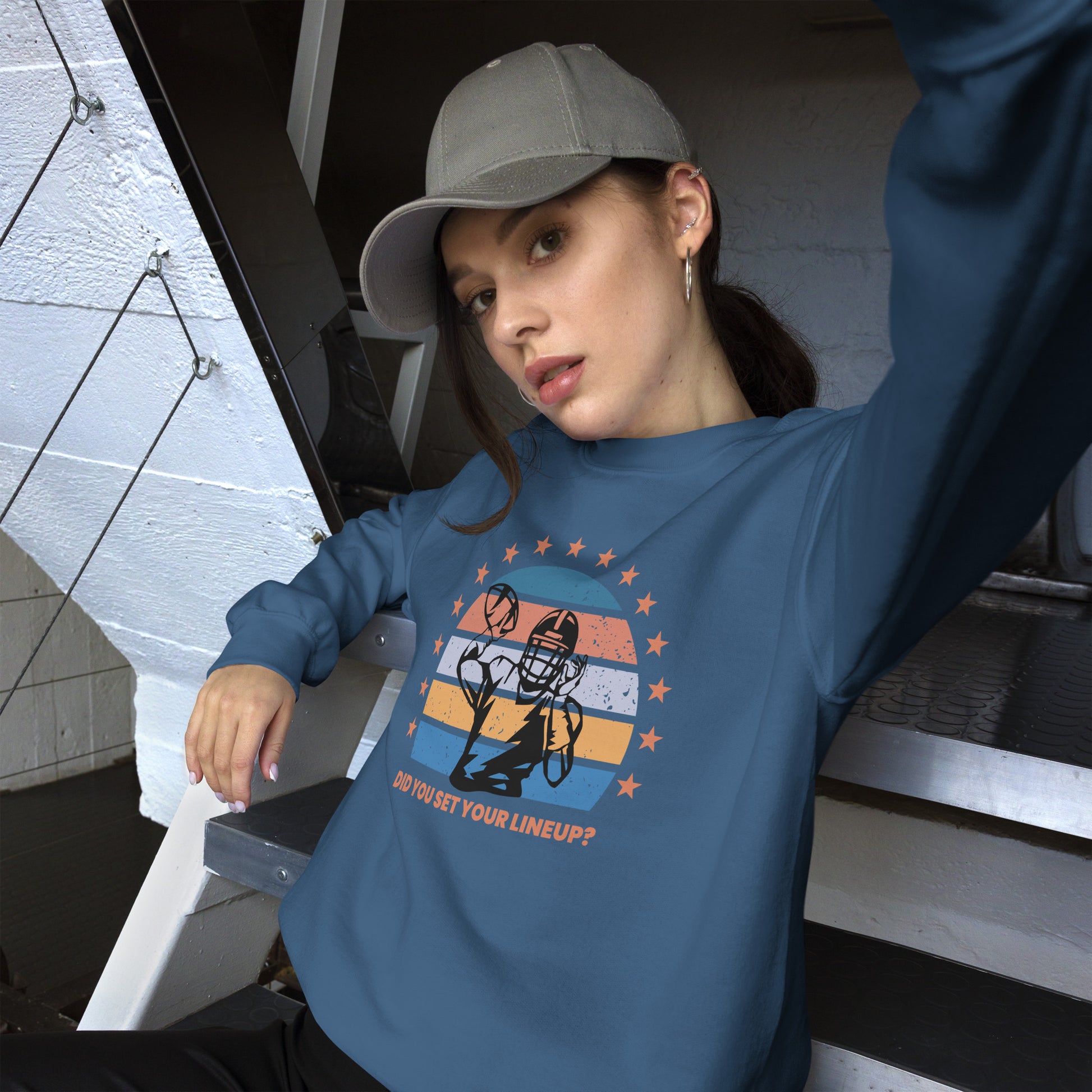 Young woman with a baseball cap wears a fantasy football sweatshirt that reads "Did you set your line up?" The sweatshirt has a silhouette of a quarterback about the throw a football with a distressed backdrop of yellow, blues, and light pink and a star border.