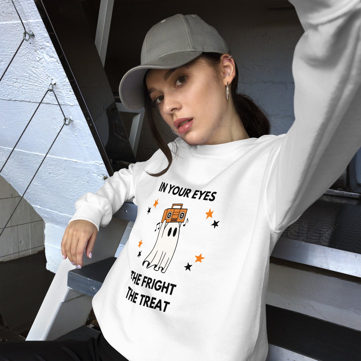 Woman in a baseball cap wears a Halloween sweatshirt featuring a ghost holding an orange boombox overhead, Say Anything style. The shirt reads "In your eyes - The Fright, The Treat."