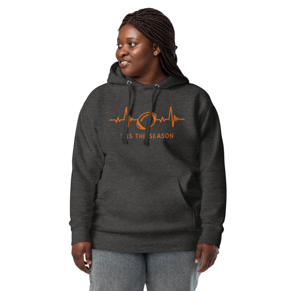 A woman with braided hair wears a dark gray football sweatshirt with orange design and text. The colors could be for the Cincinnati bengals,  Cleveland Browns, Miami Dolphins, Chicago Bears, or Denver Broncos. The charcoal gray hoodie showcases a bright orange heart monitor line with a football right in the center. The shirt reads 'TIS THE SEASON underneath.