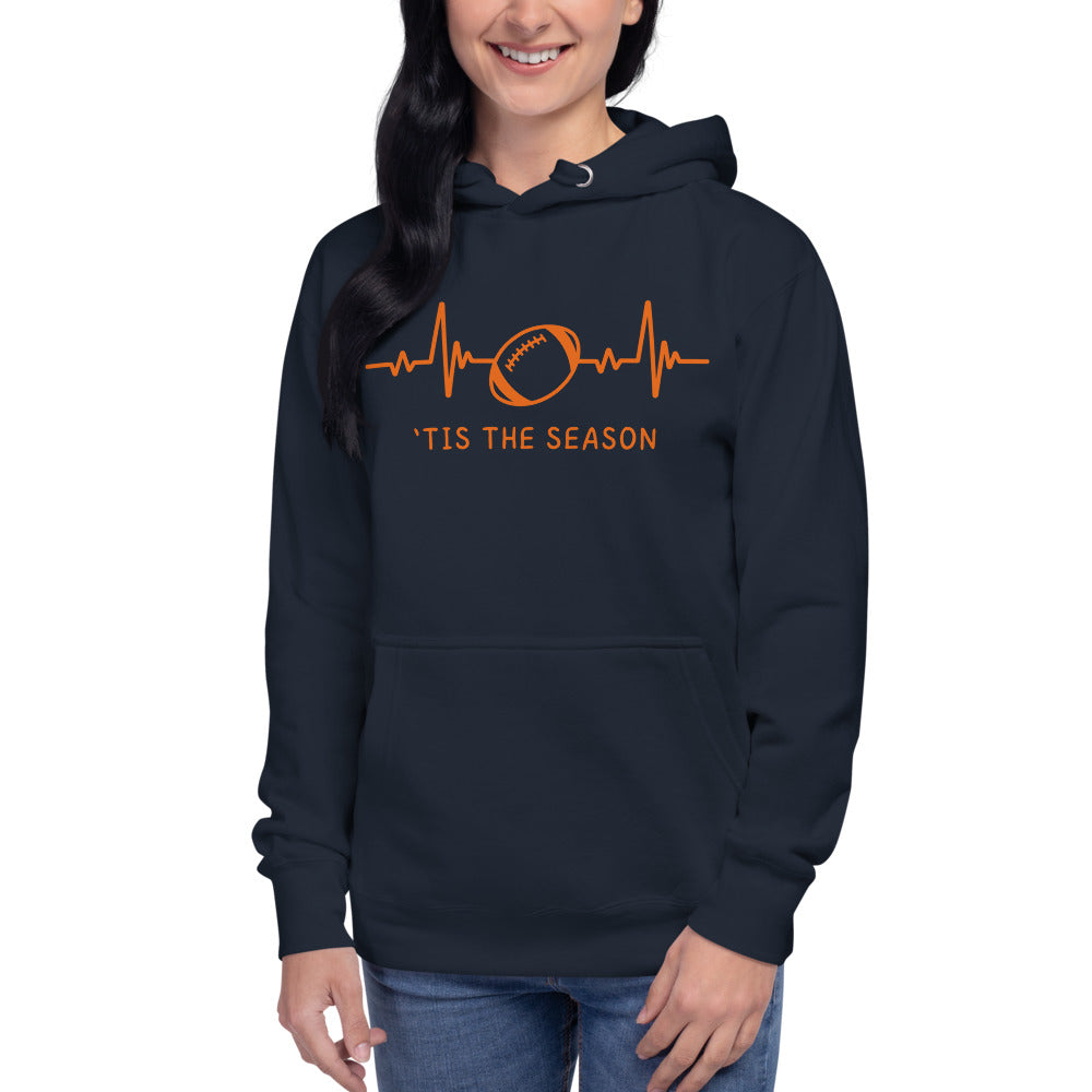 A woman with dark hair wears a football hoodie that's dark blue and orange, like the chicago bears. The navy hoodie showcases a bright orange heart monitor line with a football right in the center. The shirt reads 'TIS THE SEASON underneath.