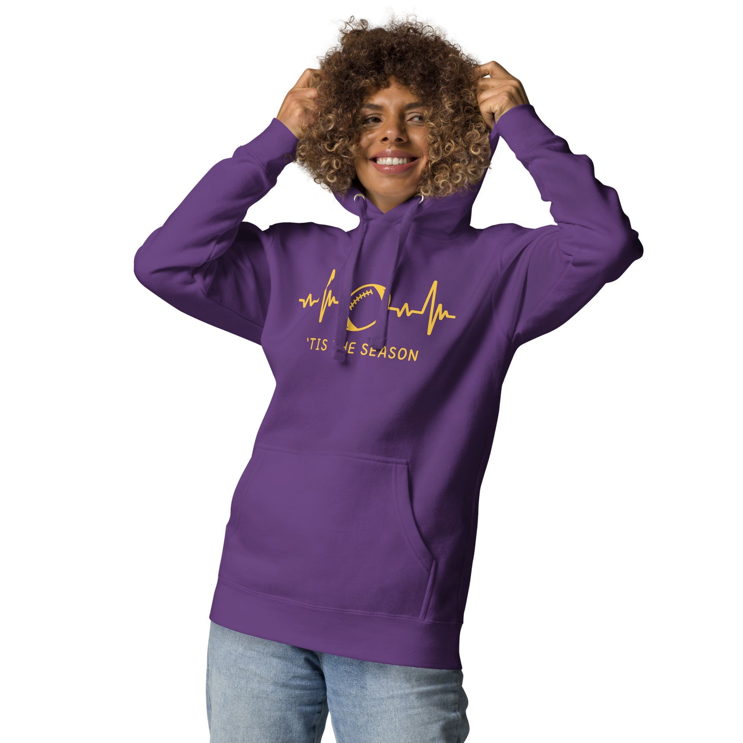 A woman with curly hair sports a purple and yellow football hoodie, looking like Minnesota Vikings colors. The purple hoodie showcases a bright yellow heart monitor line with a football right in the center. The shirt reads 'TIS THE SEASON underneath.