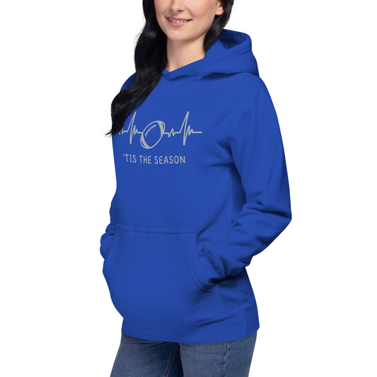 Woman wears a blue and silver-gray football sweatshirt. The bright blue hoodie showcases a silver gray heart monitor line with a football right in the center. The colors look like the Detroit Lions, Indianapolis Colts, and the Dallas Cowboys. The shirt reads 'TIS THE SEASON underneath.