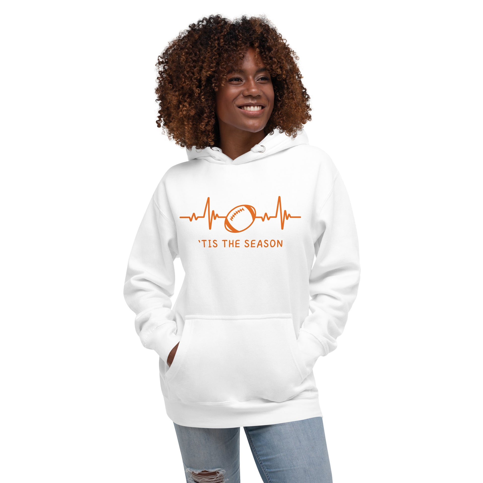 A woman with curly hair wears a white football sweatshirt with orange design and text. The colors could be for the Cincinnati bengals,  Cleveland Browns, Miami Dolphins, Chicago Bears, or Denver Broncos. The white hoodie showcases a bright orange heart monitor line with a football right in the center. The shirt reads 'TIS THE SEASON underneath.