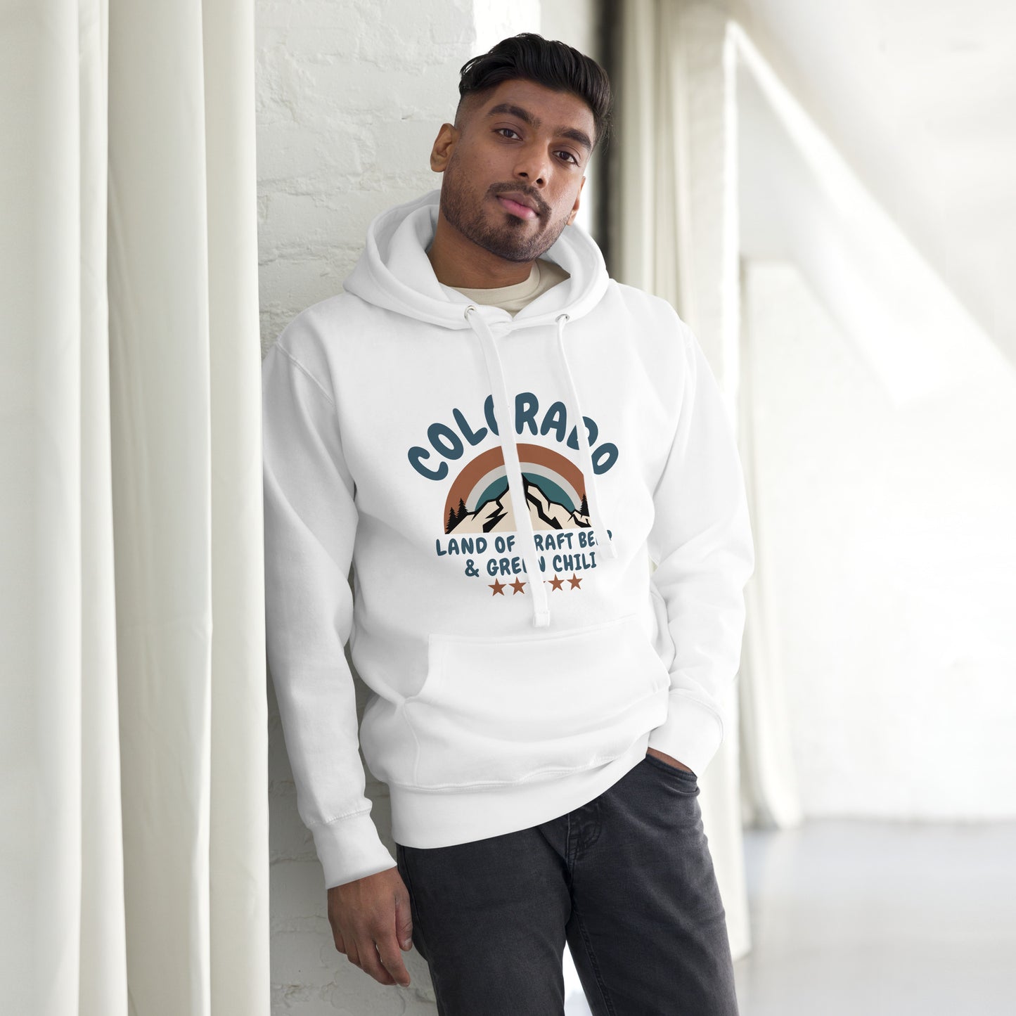 Man wearing a white hooded sweatshirt with dark jeans leans casually against a white wall. The hoodie features a Colorado-themed design with mountains, referencing craft beer and green chili, in blue and brown tones.