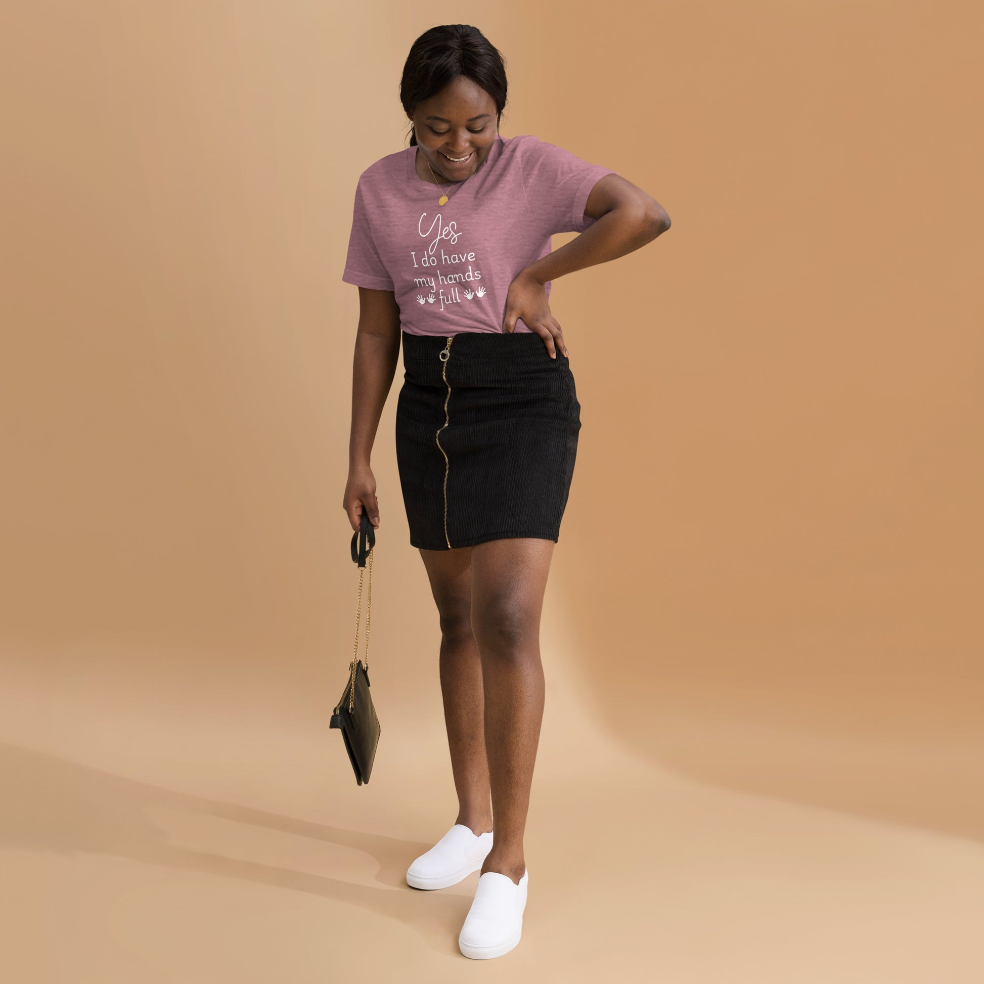 Smiling woman wears heather mauve tee that says 'Yes, I do have my hands full' with two sets of child handprints. She is also wearing a skirt and white sneakers.