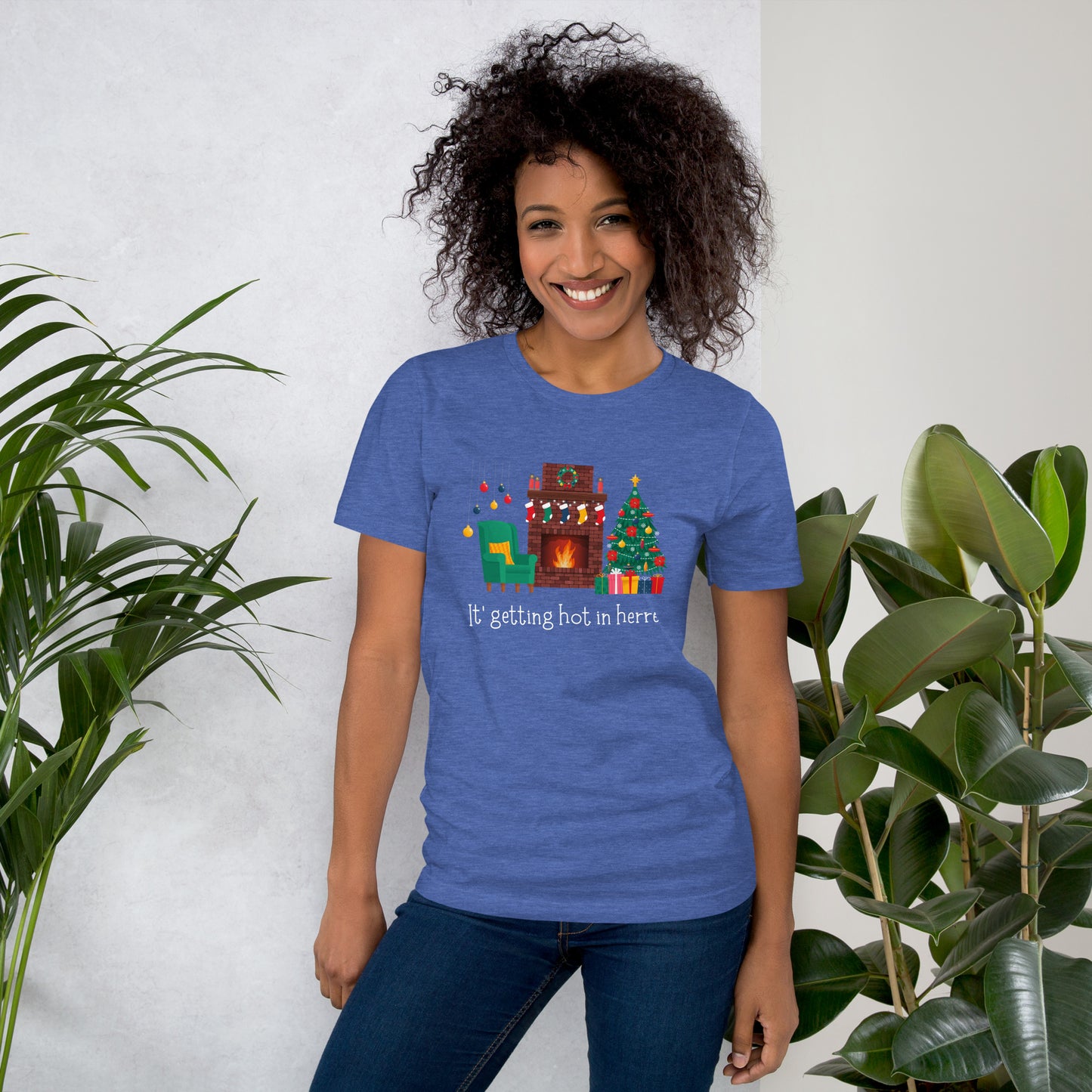 Woman wears festive holiday tee shirt that reads "It's getting hot in herre". The design is a cozy Christmas living room with holiday decorations and a roaring fireplace.