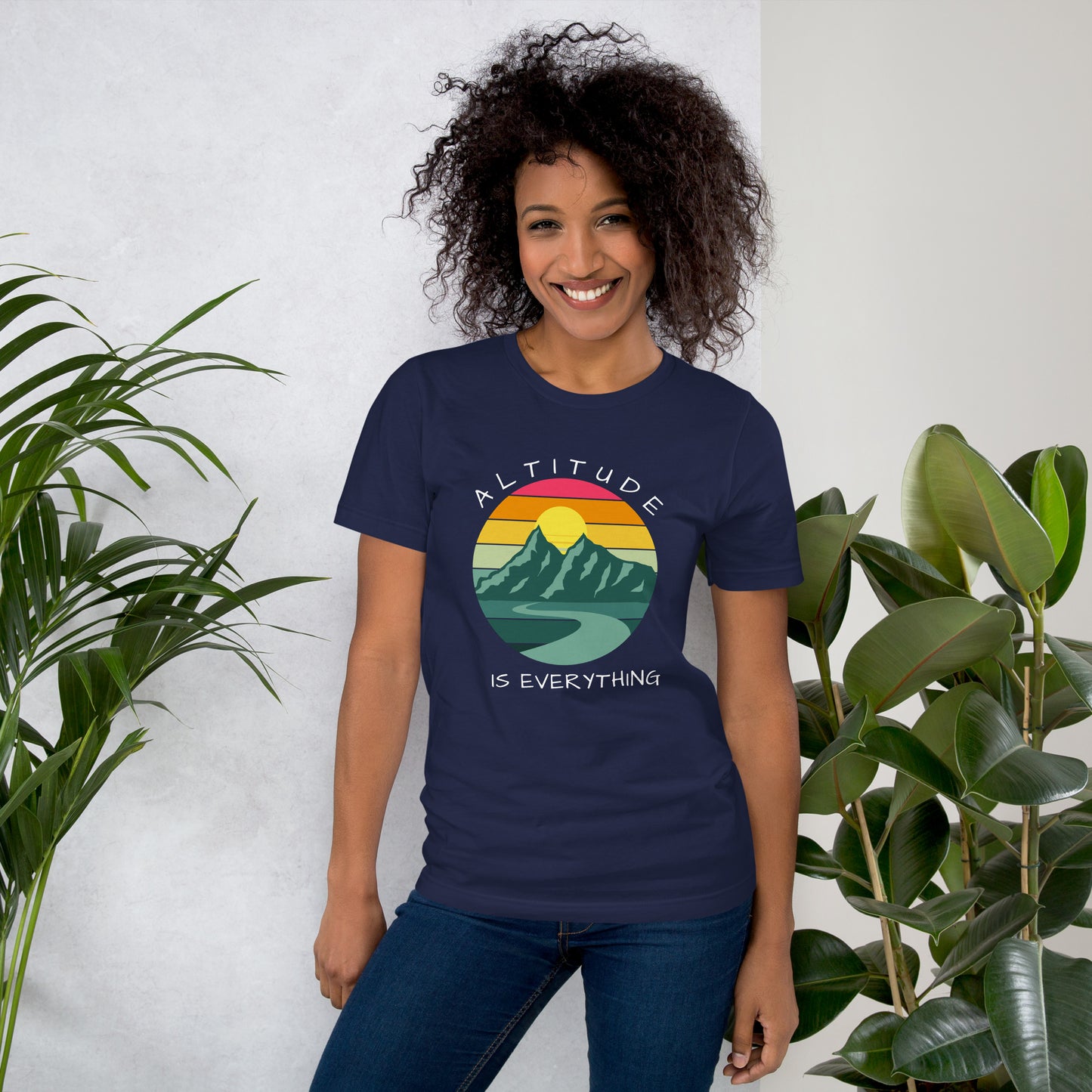 Smiling woman with curly hair wears jeans and a vivid navy graphic tee that reads "ALTITUDE IS EVERYTHING." The image is a beautiful mountain scene with a colorful sunset. 