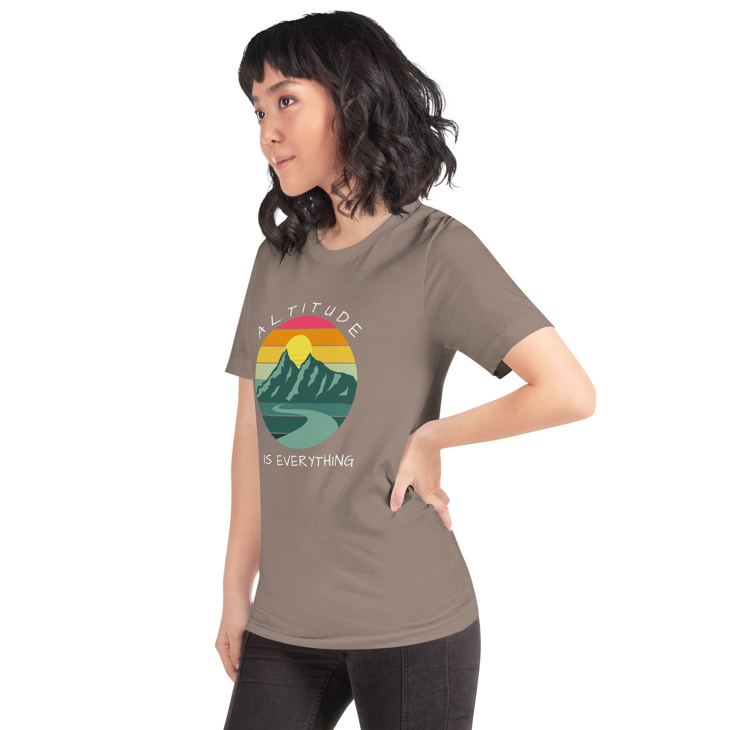 Woman with dark hair wears jeans and a pebble brown graphic tee that reads "ALTITUDE IS EVERYTHING." The image is a beautiful mountain scene with a colorful sunset. 