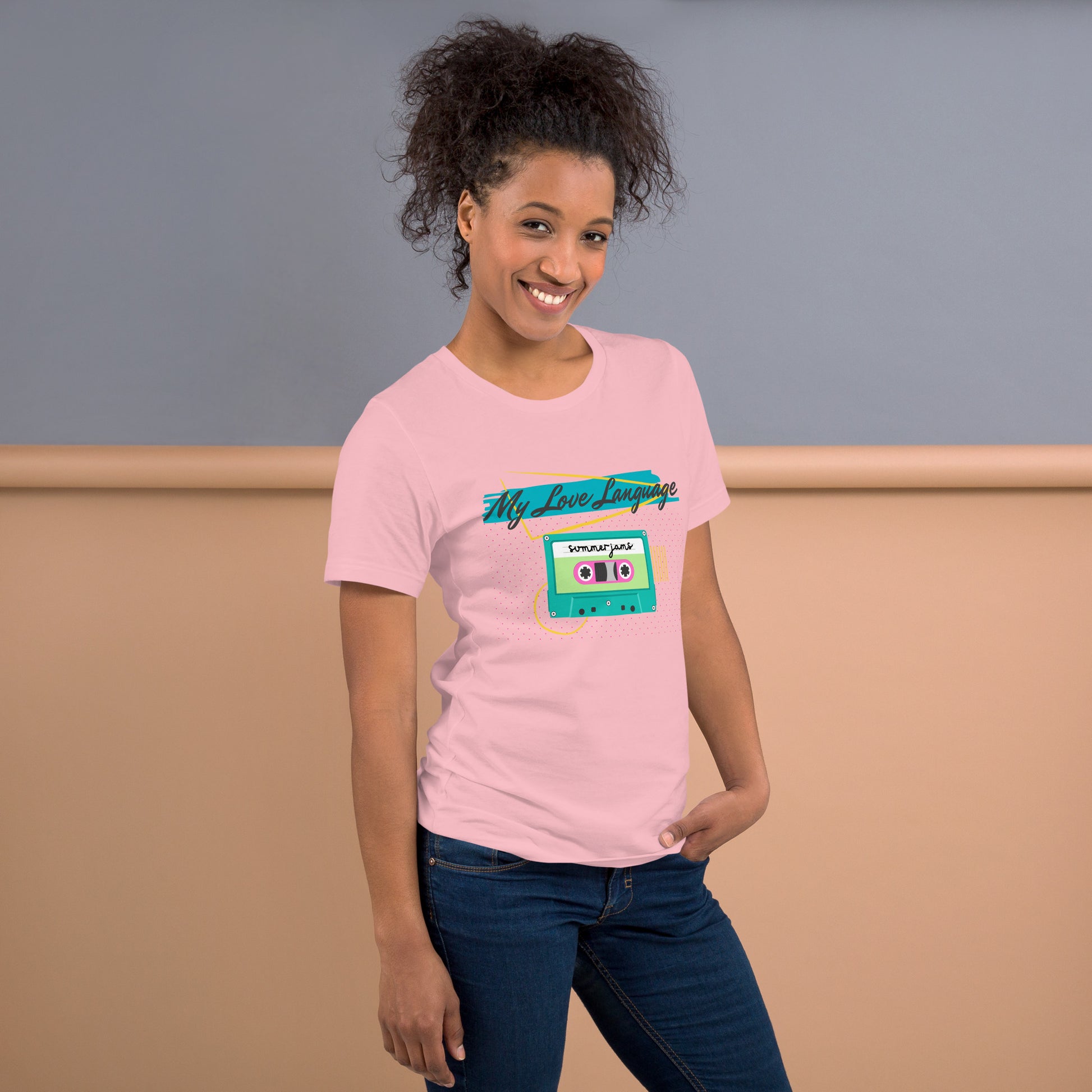 A woman wears a soft pink tee with bright pink, blue, yellow and green graphics from the 80s and 90s. The shirt reads "MY LOVE LANGUAGE" and features a cassette tape - a reference to the mixtape era.