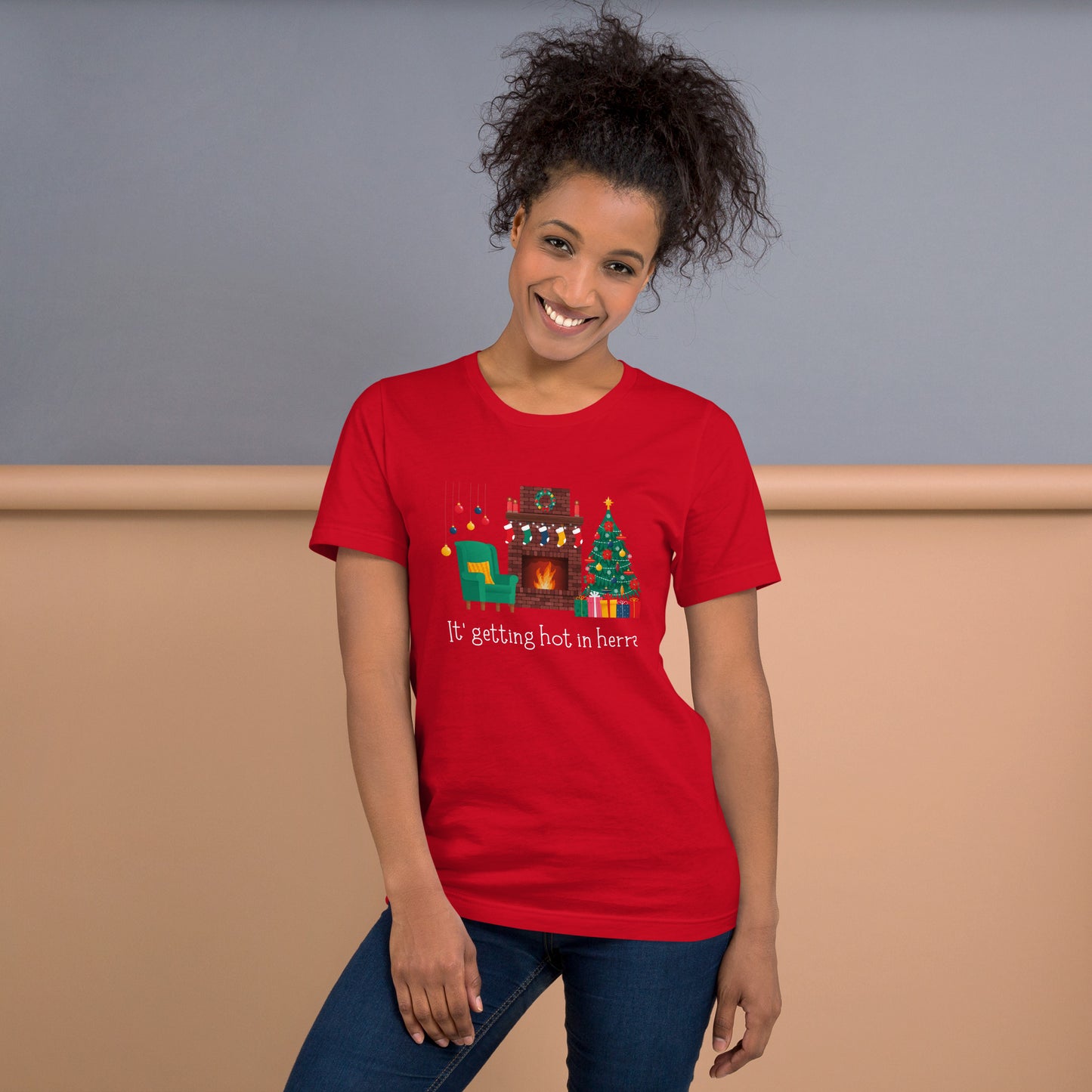 Woman wears festive red holiday tee shirt that reads "It's getting hot in herre". The design is a cozy Christmas living room with holiday decorations and a roaring fireplace.