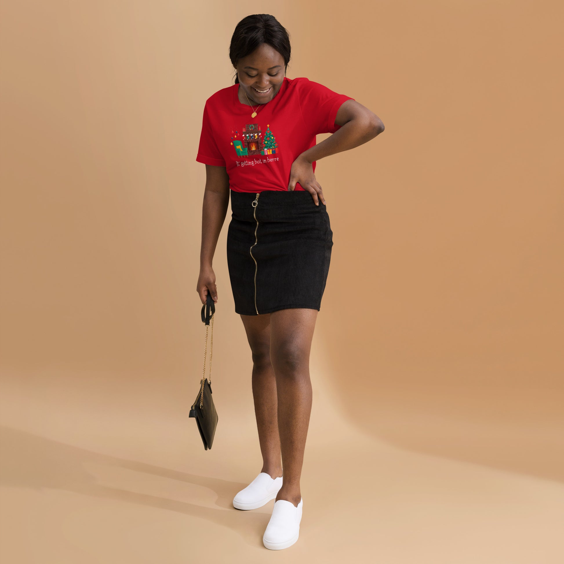 Woman wears festive red holiday tee shirt that reads "It's getting hot in herre". The design is a cozy Christmas living room with holiday decorations and a roaring fireplace.