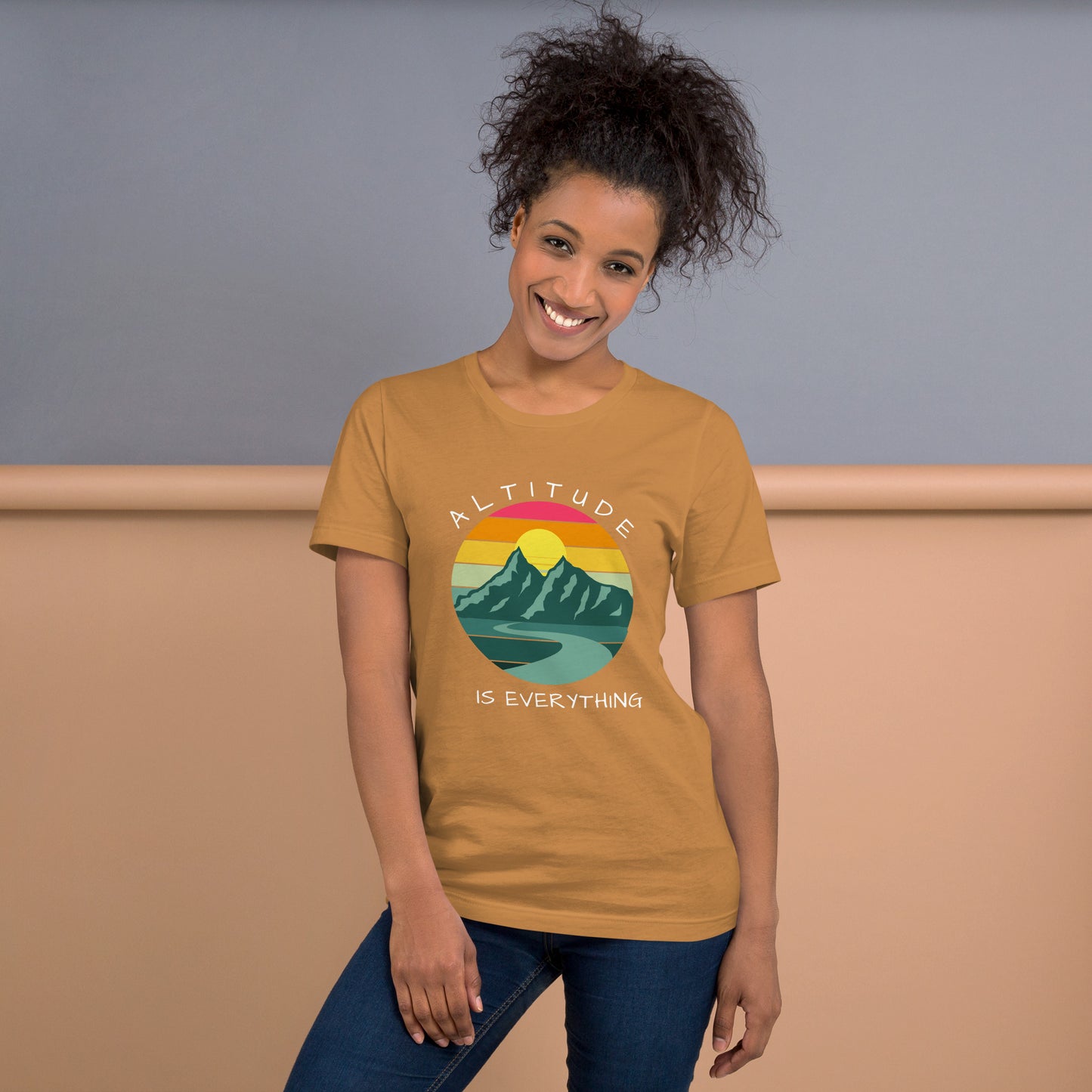 Smiling woman with curly hair wears a tan brown graphic tee that reads "ALTITUDE IS EVERYTHING." The image is a beautiful mountain scene with a colorful sunset. 