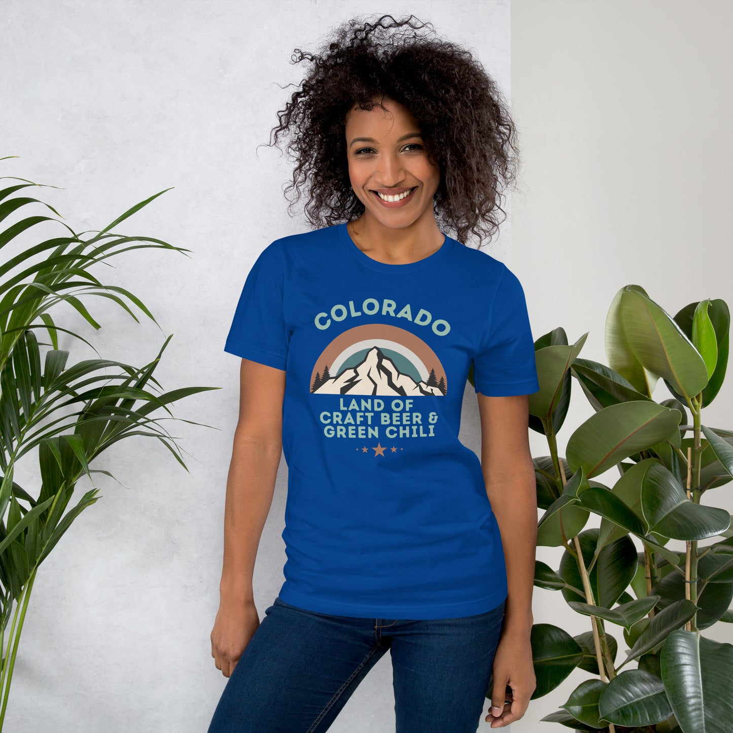 Smiling woman with curly dark hair wears a graphic tee that says "COLORADO: Land of Craft Beer & Green Chili".  The shirt includes a scenic view of the mountains with a colorful sky.