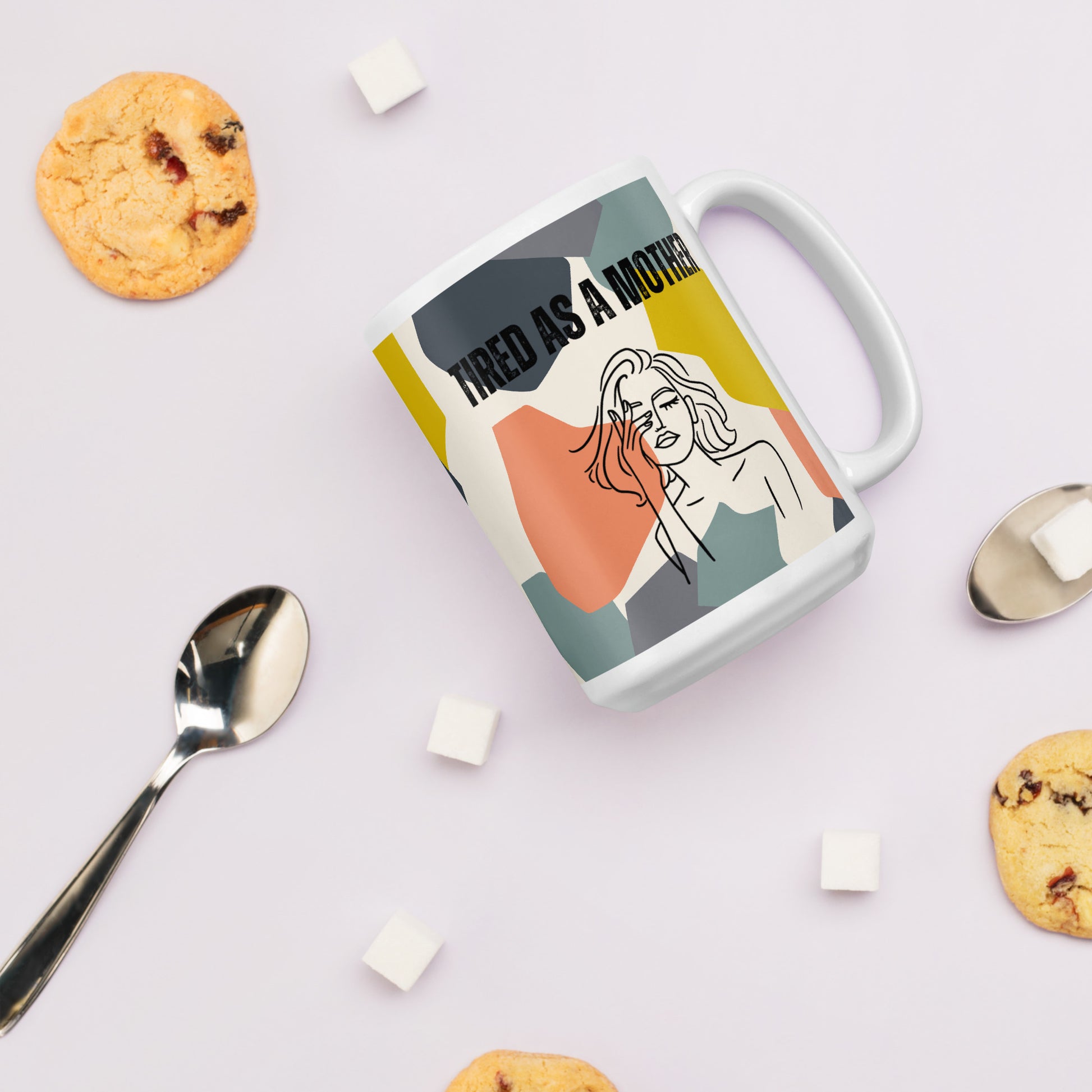 Colorful "tired and a mother" mug sits on a counter amidst a couple of spoons, several marshmallows, and cookies. 