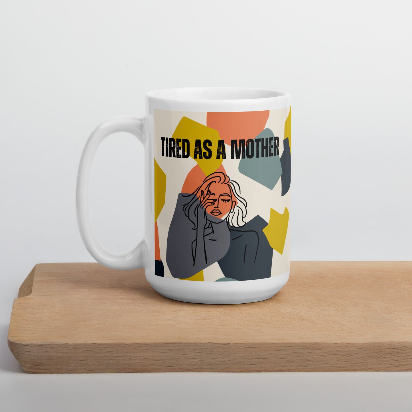 Colorful ceramic mug sits on a wooden board. white ceramic mug featuring the text "TIRED AS A MOTHER" and an abstract illustration of a weary woman with tousled hair. The design incorporates a modern, colorful geometric background with shapes in yellow, orange, gray, and teal.