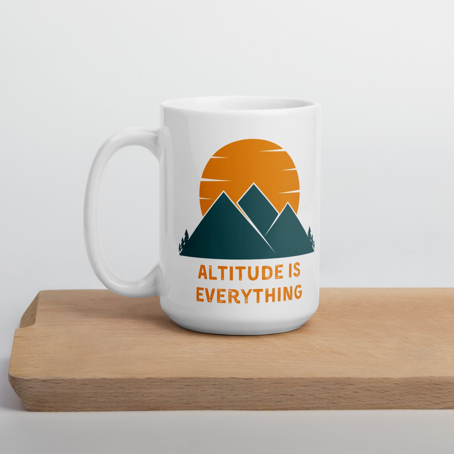 White ceramic mug that says Altitude is Everything sits on a wooden shelf. The mug displays a  graphic image of mountains and a sunset in orange and deep greenish-blue.