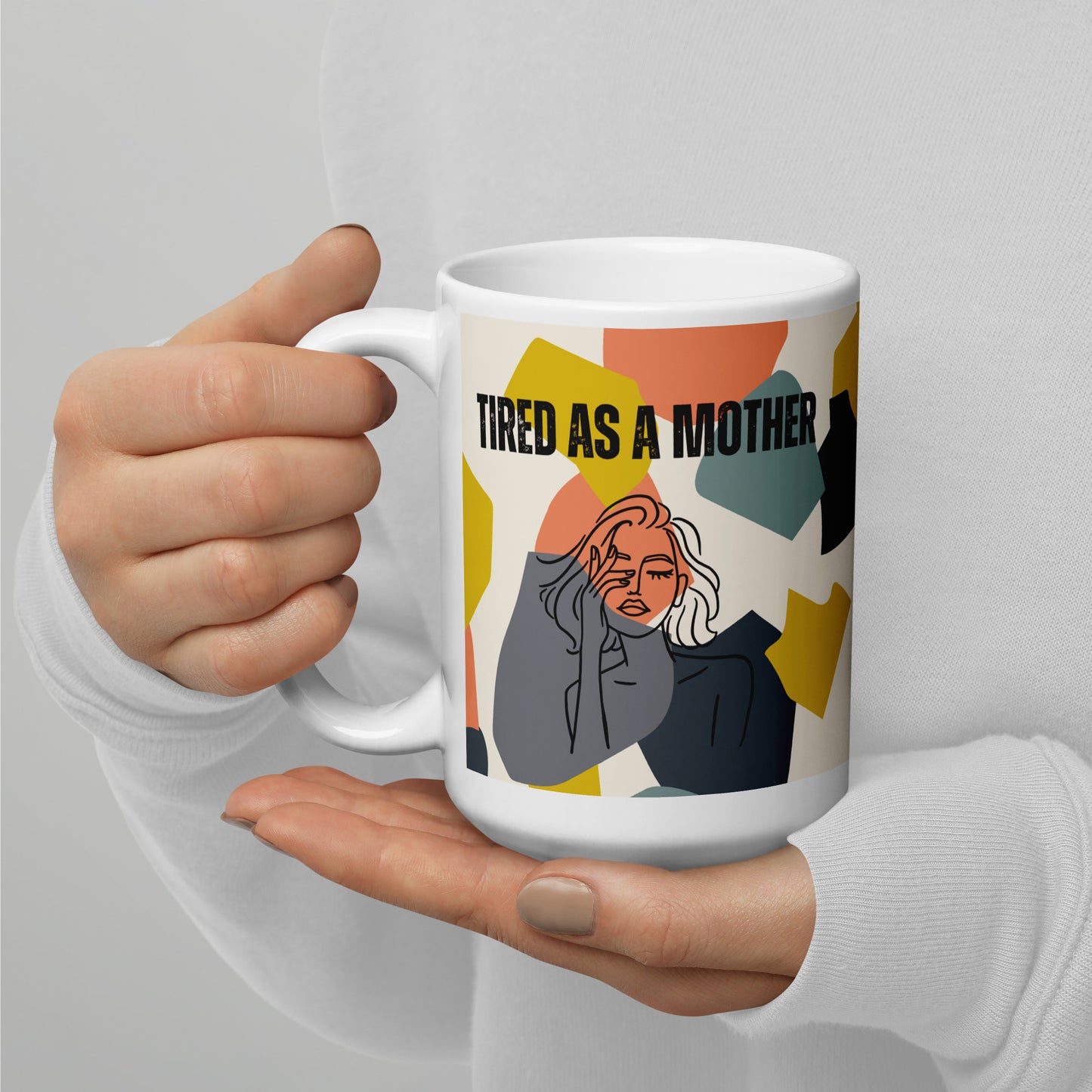 Manicured hands cup a White ceramic mug with "TIRED AS A MOTHER" text and stylized illustration of a weary woman, set against a colorful geometric background. 