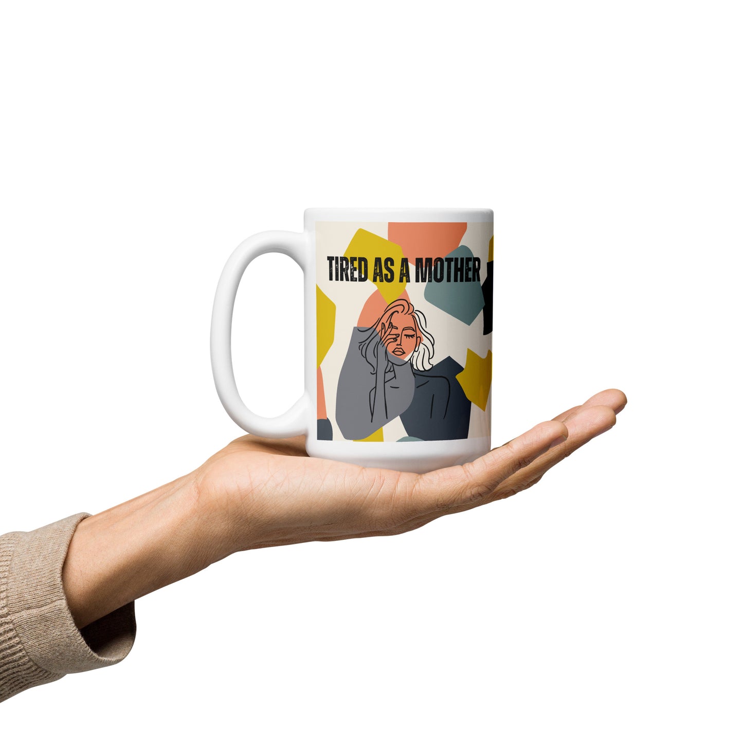 Outstretched hand holds a 15 oz white ceramic mom mug - white ceramic mug featuring the text "TIRED AS A MOTHER" and an abstract illustration of a weary woman with tousled hair. The design incorporates a modern, colorful geometric background with shapes in yellow, orange, gray, and teal.