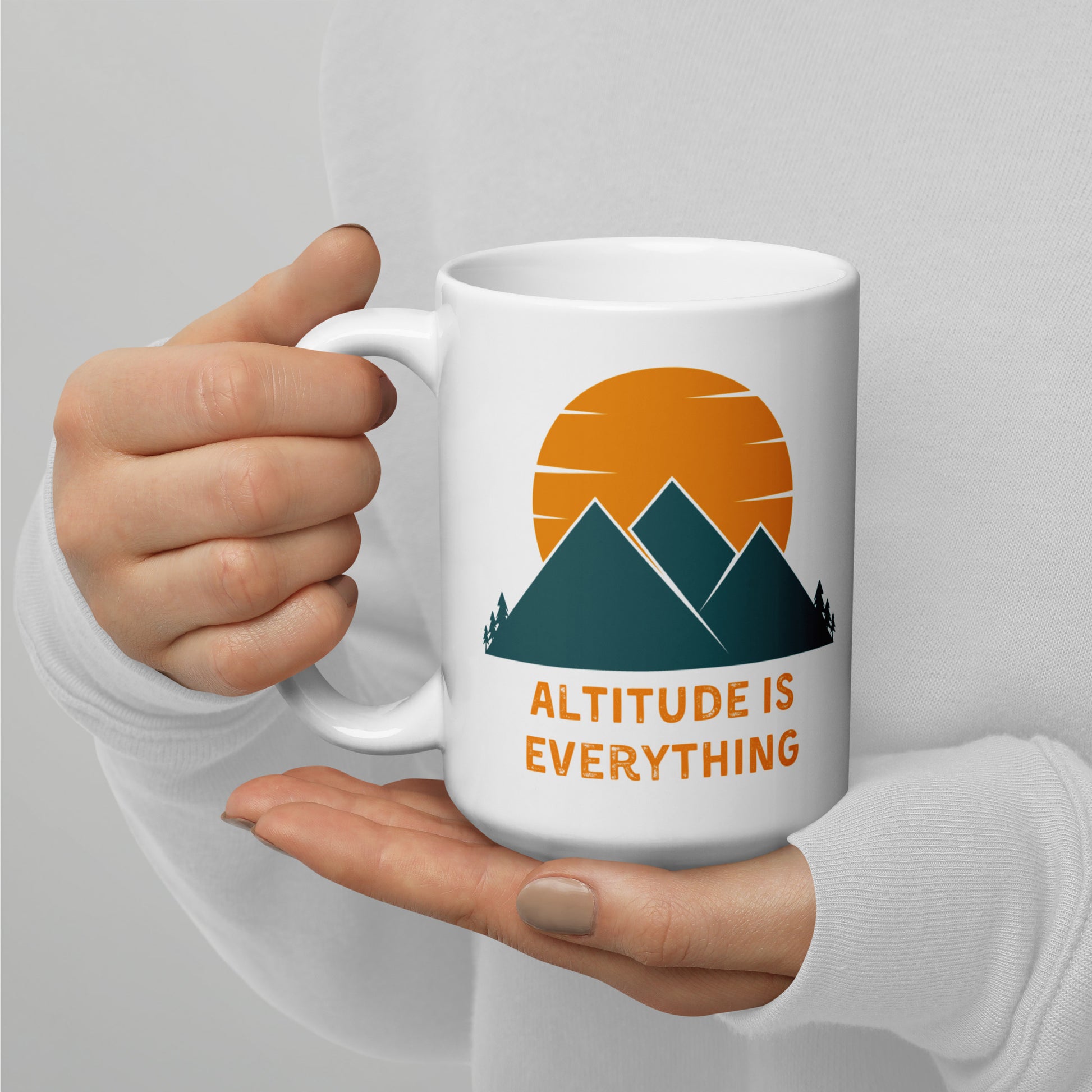 The image shows a white ceramic mug with a graphic design of mountains and a sunset, along with the text "ALTITUDE IS EVERYTHING", captured in the hands of a person.